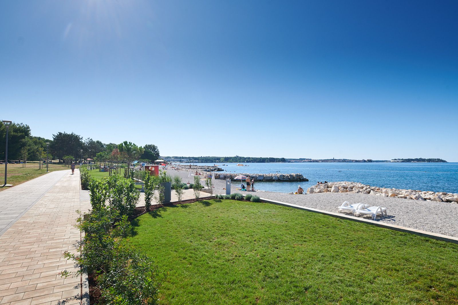 Ein sonniger Küstenweg mit Grünflächen, Strand und ruhigem Wasser, ideal für einen entspannten Tag am Meer.