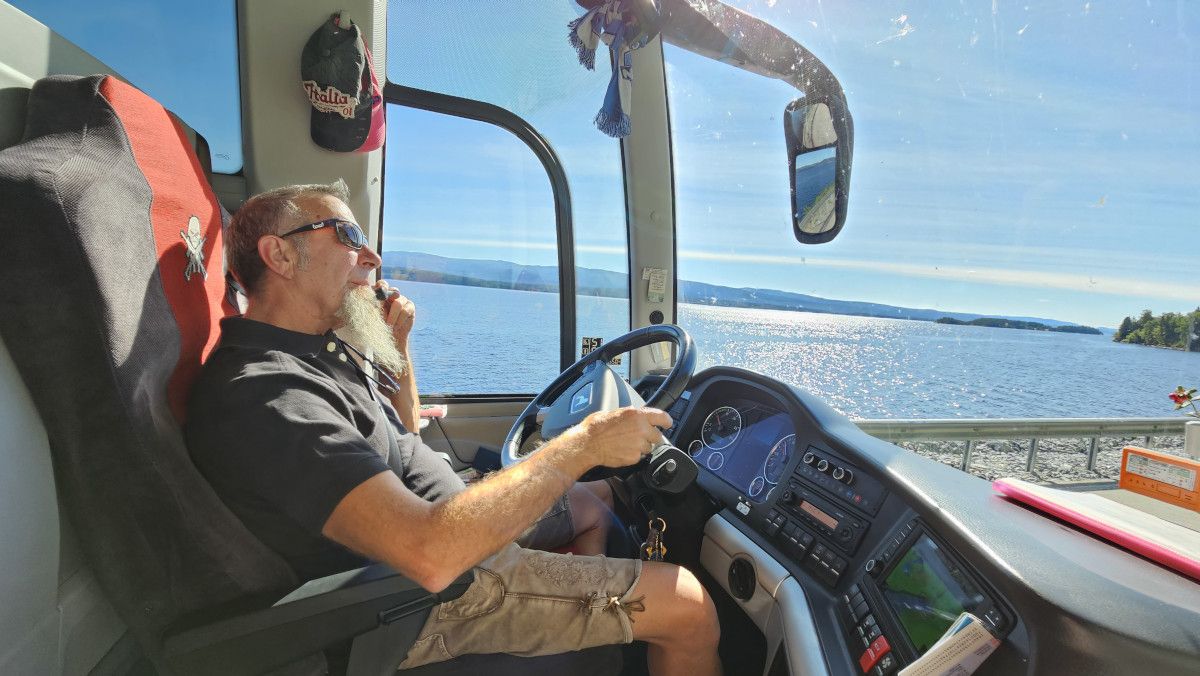 Ein Mann mit Bart fährt einen Bus in der Nähe eines glitzernden Sees. Sonne und blauer Himmel sind sichtbar.