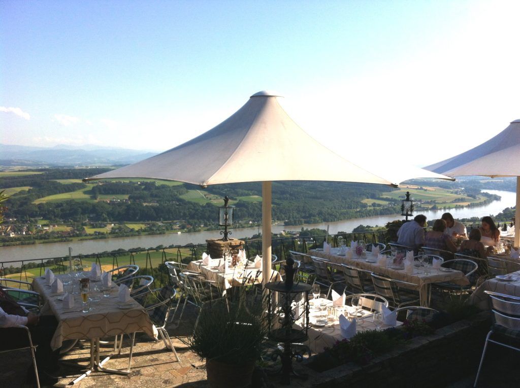 Eine Terrasse mit Tischgedecken, Sonnenschirmen und Blick auf einen Fluss und die umliegende Landschaft.