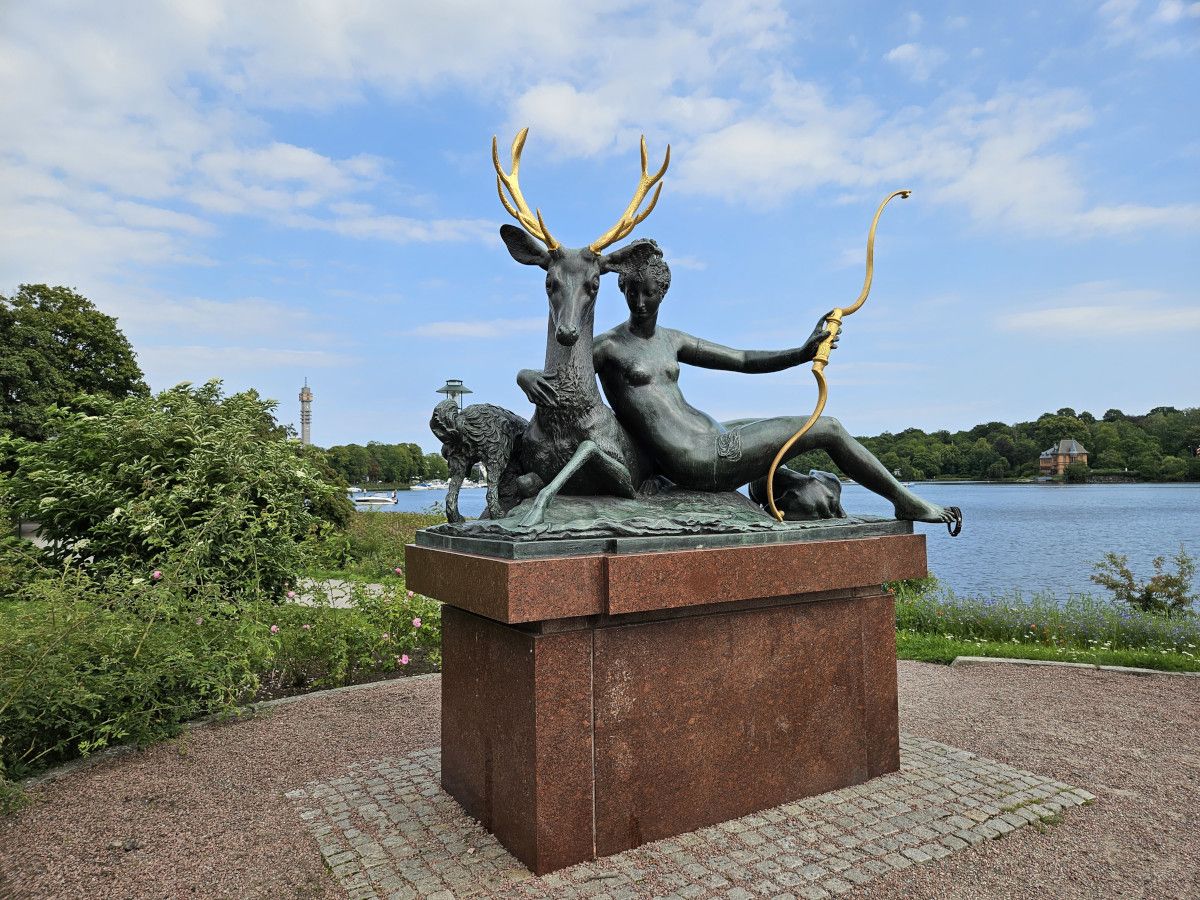 Eine Statue zeigt eine Frau mit einem Hirsch und einem Hund, am Wasser, umgeben von grünem Park.