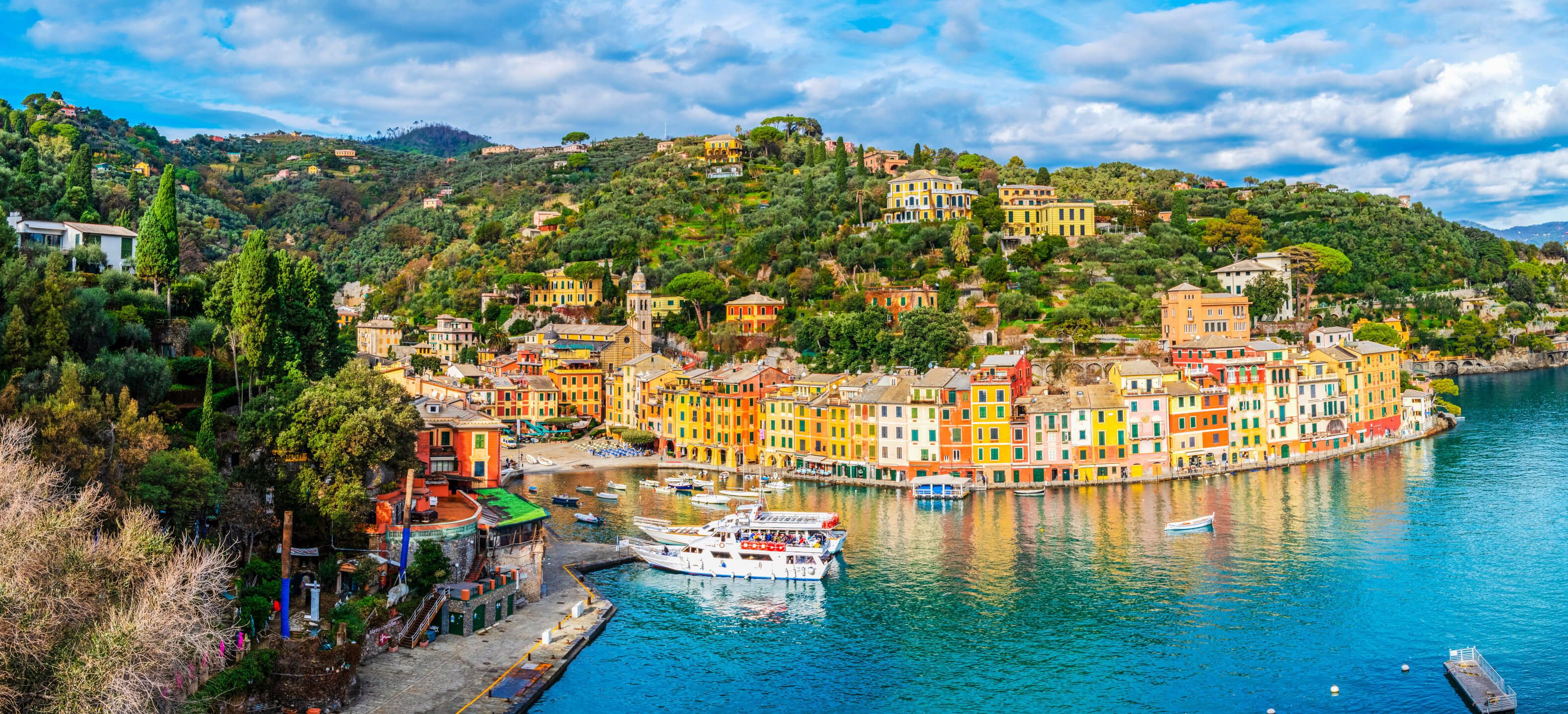 Bunte Häuser am Hafen von Portofino, umgeben von grünen Hügeln und klarem Wasser unter einem bewölkten Himmel.