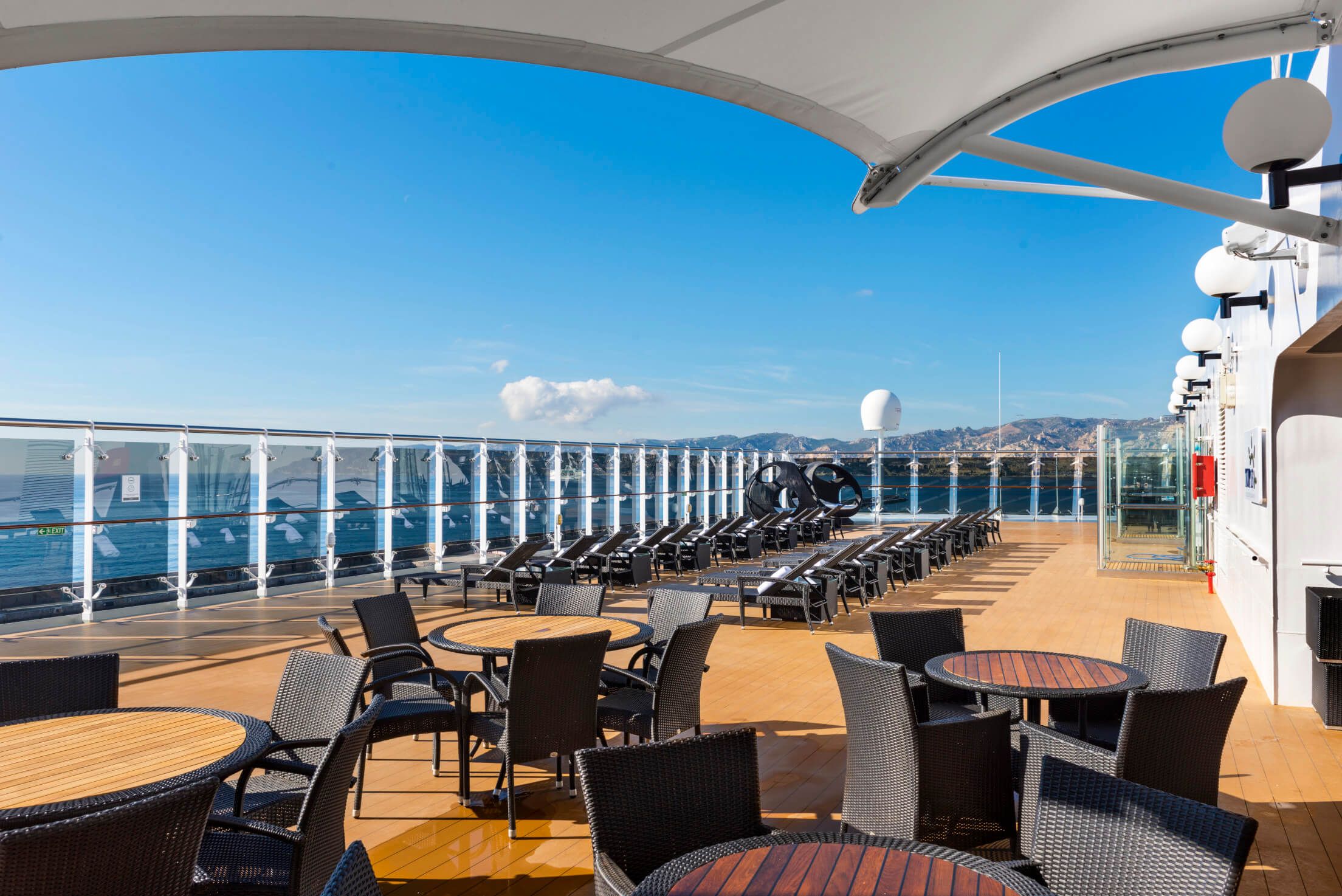 Ein sonniger Außendeckbereich eines Schiffs mit Lounge-Möbeln und Blick auf das Wasser.