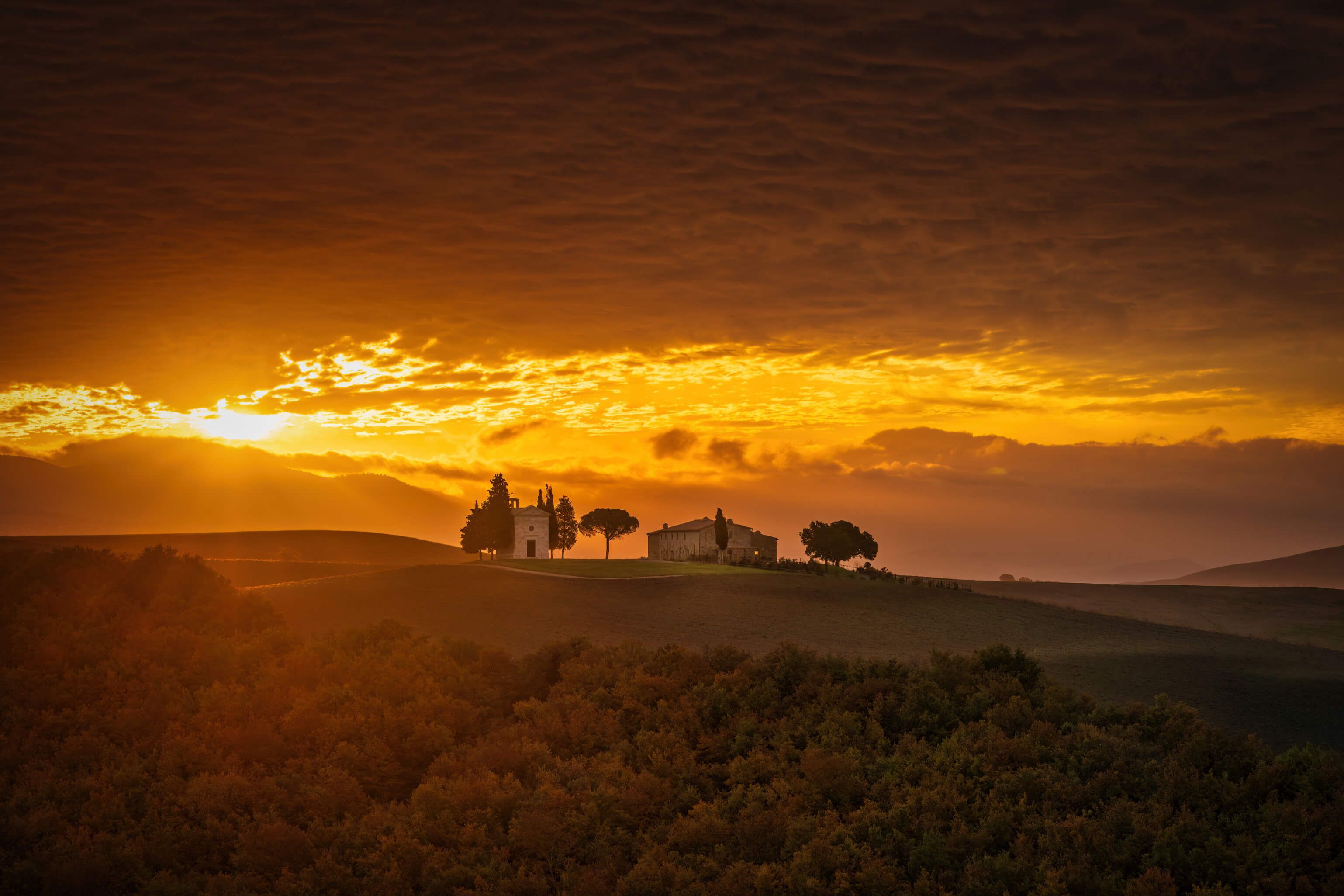 Eine malerische Landschaft mit goldenem Sonnenuntergang, sanften Hügeln und verstreuten Bäumen und Gebäuden.