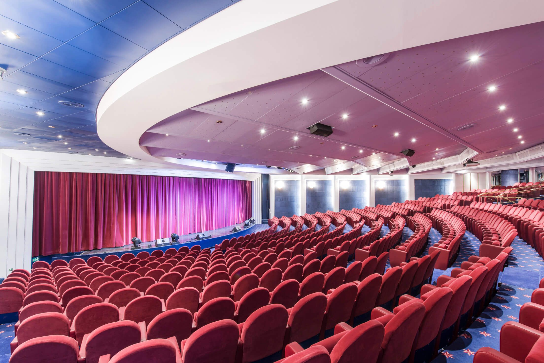 Ein modernes Theater mit roten Sitzen, einer Bühne und einem lila Vorhang. Gemütliche Atmosphäre.