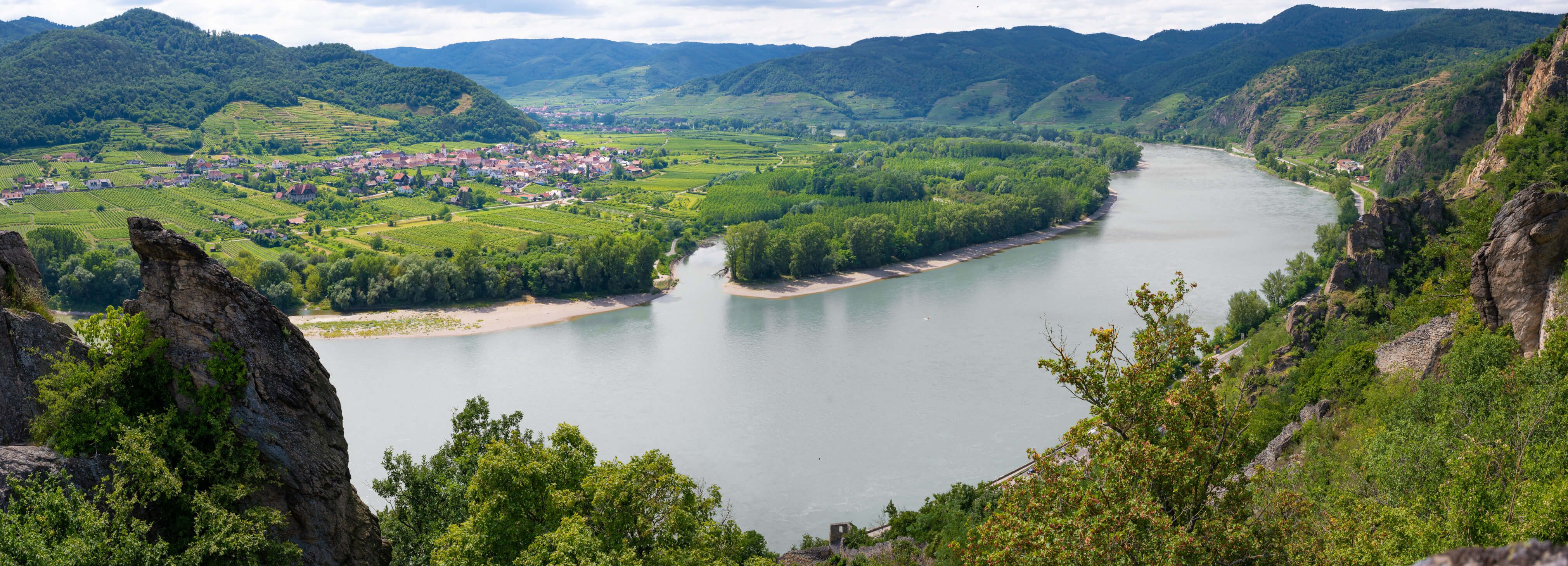 Eine malerische Landschaft mit einem Fluss, Weinbergen und sanften Hügeln unter einem bewölkten Himmel.