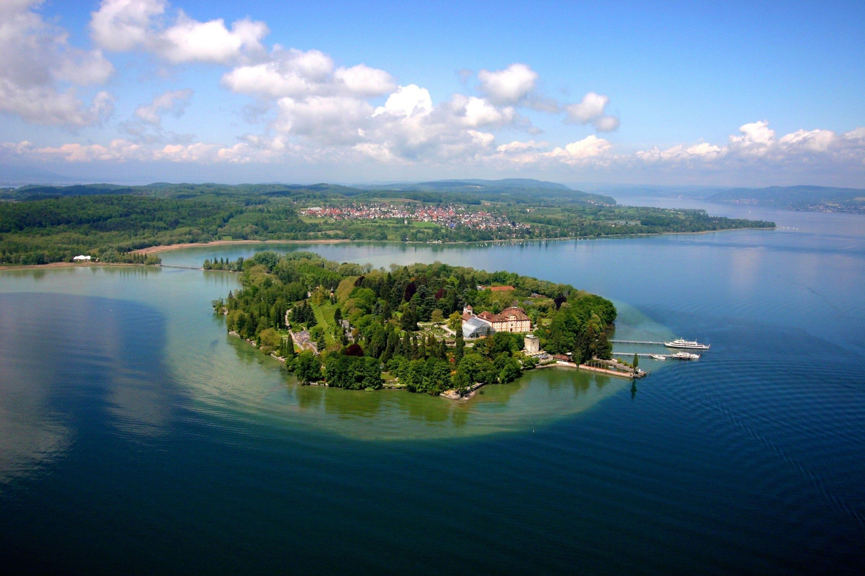 Die grüne Insel  Mainau im Bodensee im blauen Wasser, umgeben von Wäldern und sanften Hügeln unter einem wolkigen Himmel.