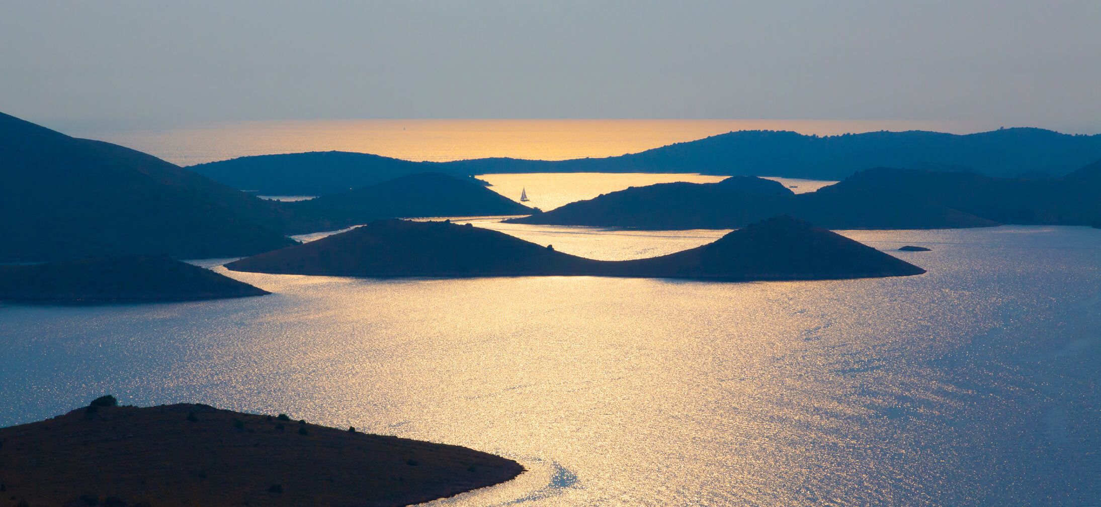 Eine ruhige Landschaft mit sanften Hügeln, glitzerndem Wasser und einem Segelboot in der Ferne.
