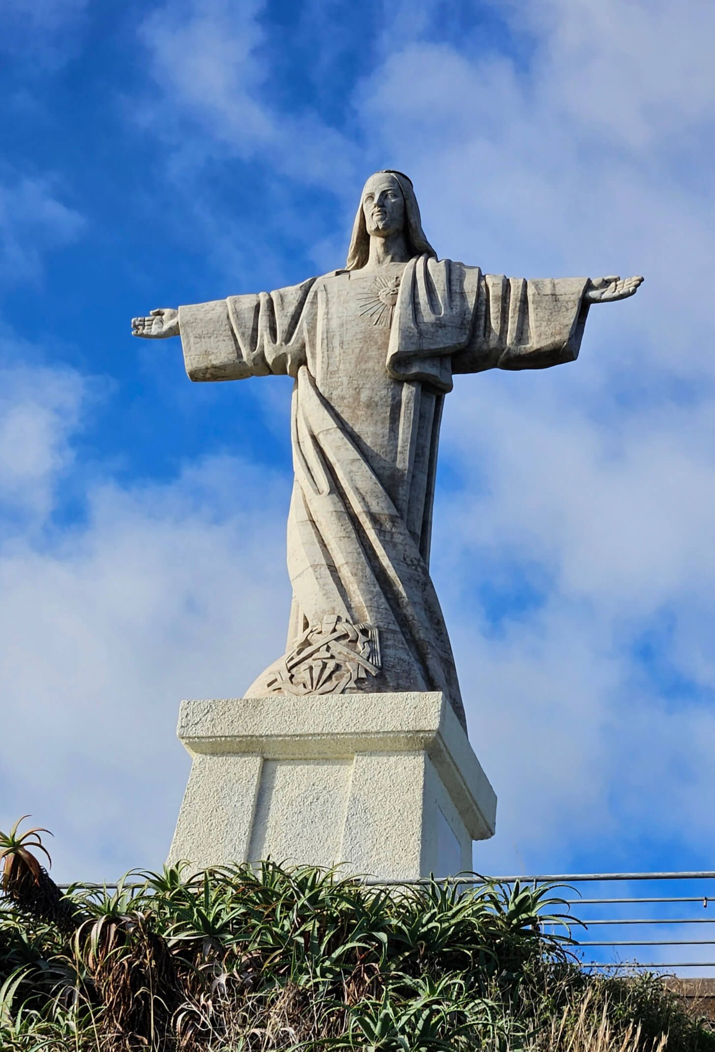 Eine majestätische Statue Jesu mit ausgestreckten Armen, vor einem blauen Himmel und grüner Vegetation.