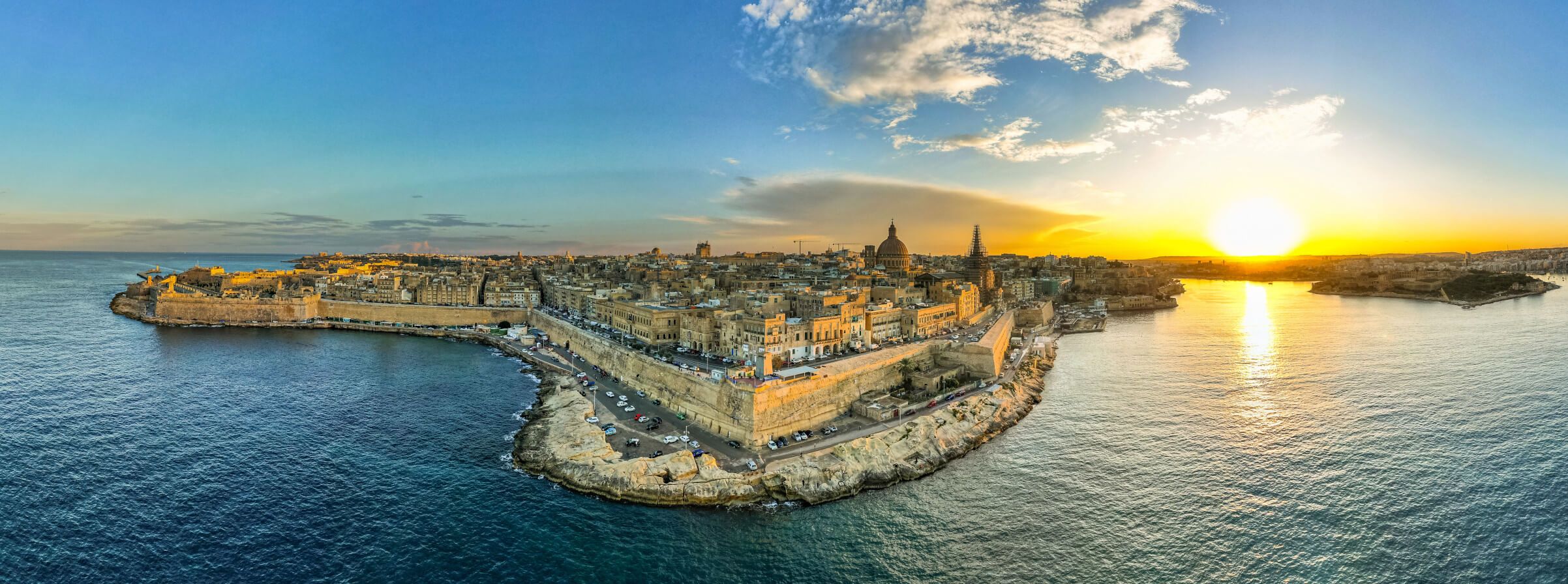 Malta bei Sonnenuntergang: Die Altstadt und die Küste spiegeln goldenes Licht im ruhigen Wasser wider.