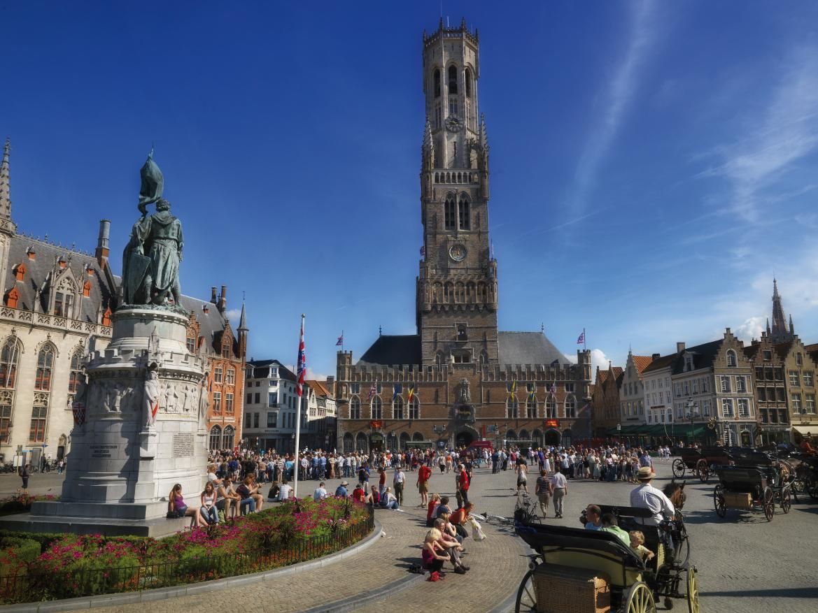 Der Platz mit dem beeindruckenden Turm, Statuen und vielen Menschen vor historischen Gebäuden in strahlendem Sonnenschein.