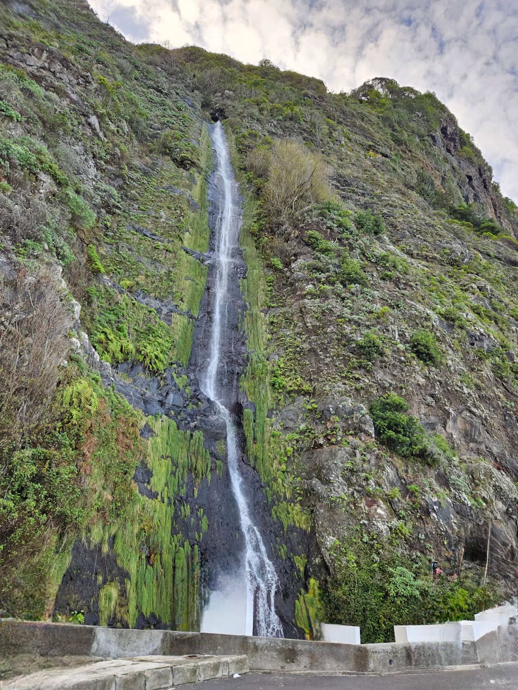 Ein beeindruckender Wasserfall fließt über bewachsene Felsen in eine grüne, natürliche Landschaft.
