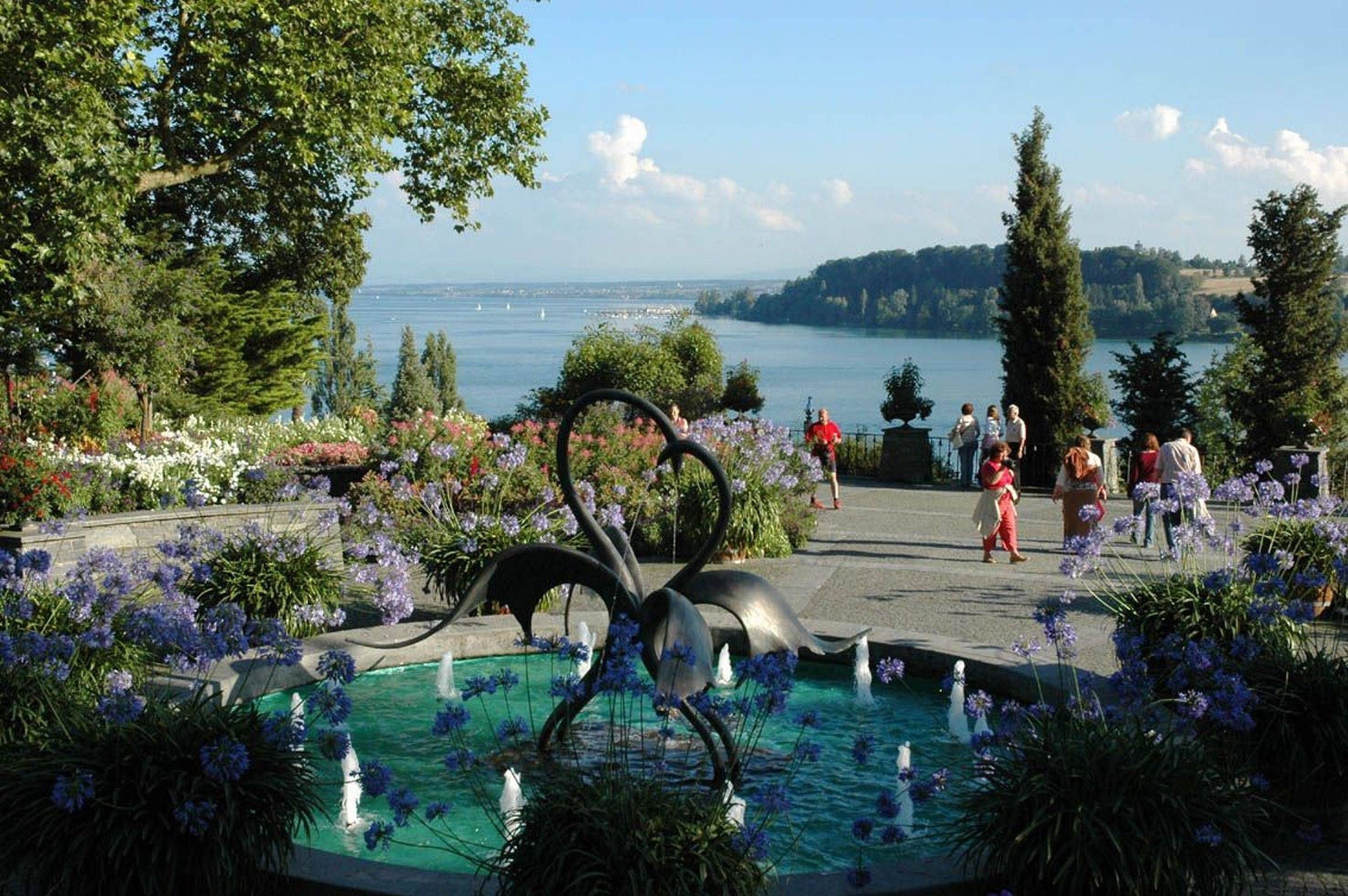 Ein schöner Garten mit Blumen, einem Brunnen und Blick auf den See, während Menschen spazieren gehen.