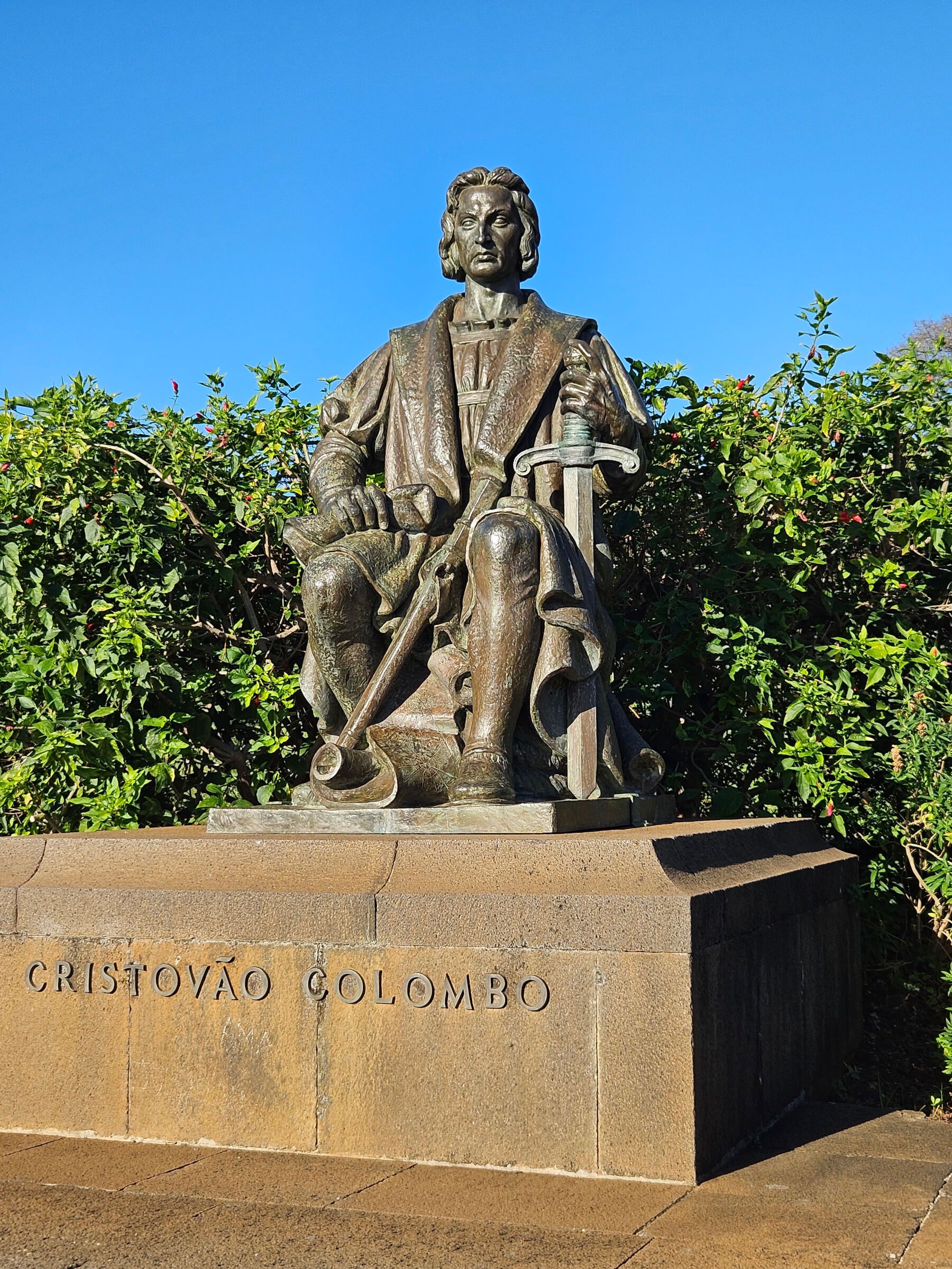 Eine Statue von Christoph Kolumbus sitzt auf einem Sockel, umgeben von grünen Pflanzen und blauem Himmel.