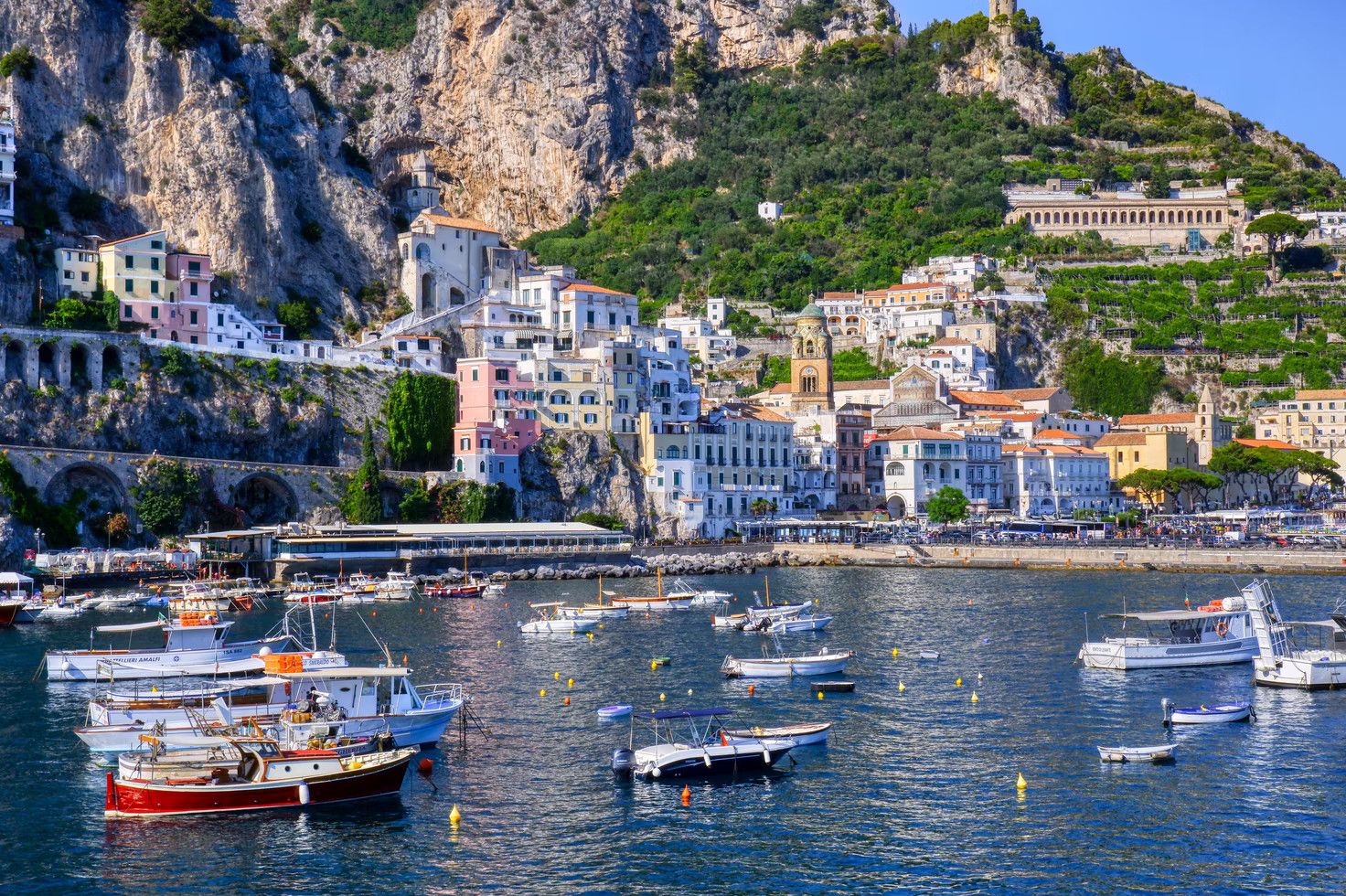 Bunte Boote im Wasser, umgeben von malerischen Häusern an einer bewaldeten Küste. Ein sonniger Tag in Italien.