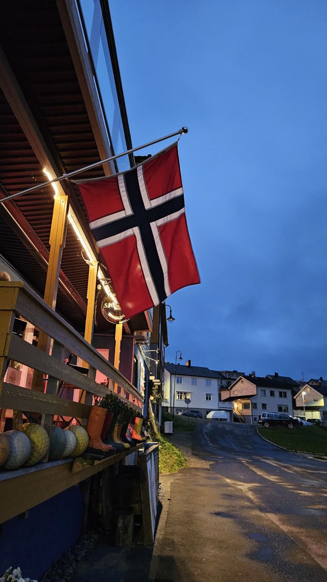 Norwegische Flagge weht an einem Gebäude bei Dämmerung, mit bunten Gummistiefeln und Häusern im Hintergrund.