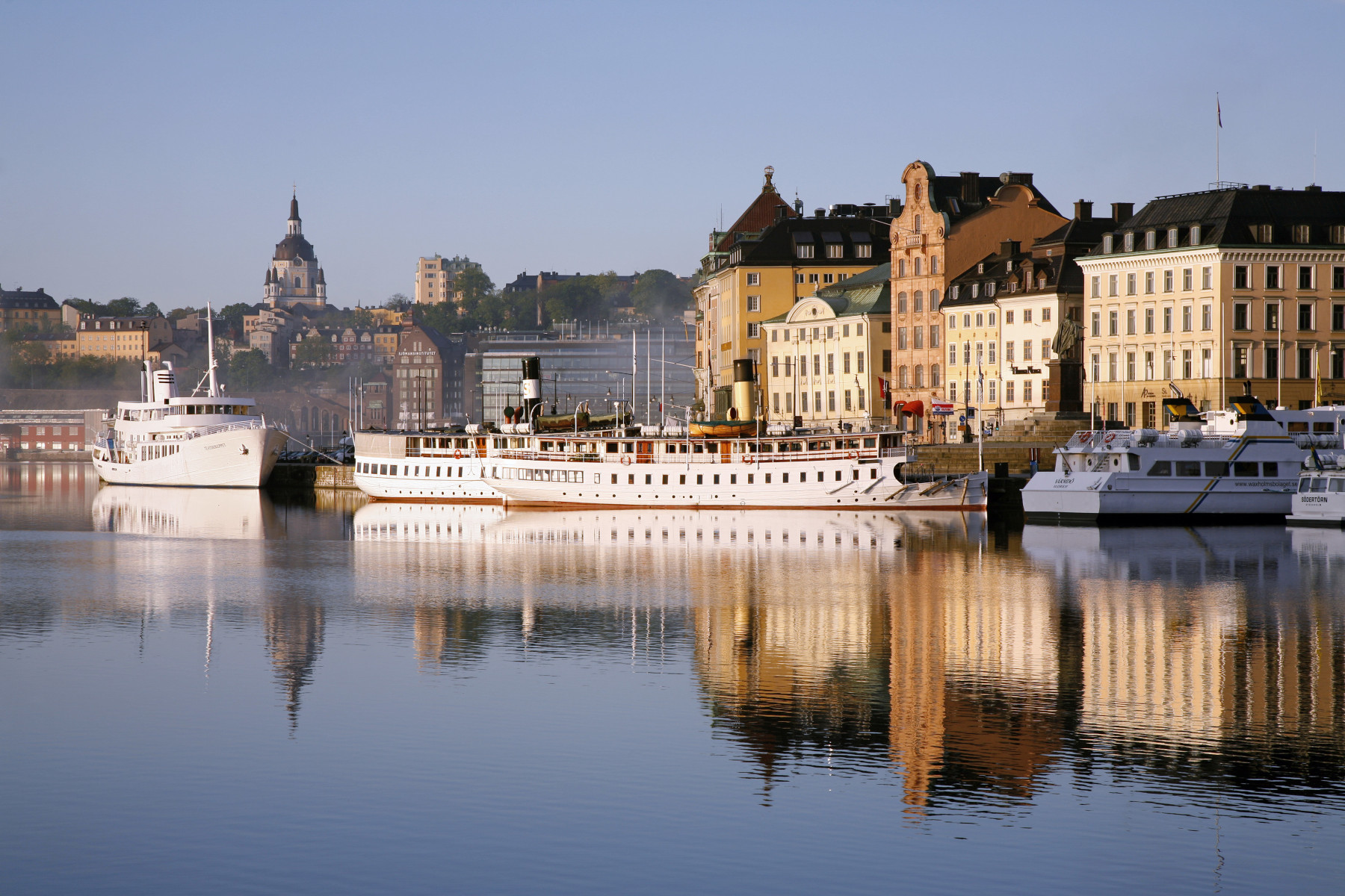 Ein malerischer Hafen mit Booten und reflektierenden Gebäuden im Hintergrund bei ruhigem Wasser und blauem Himmel.