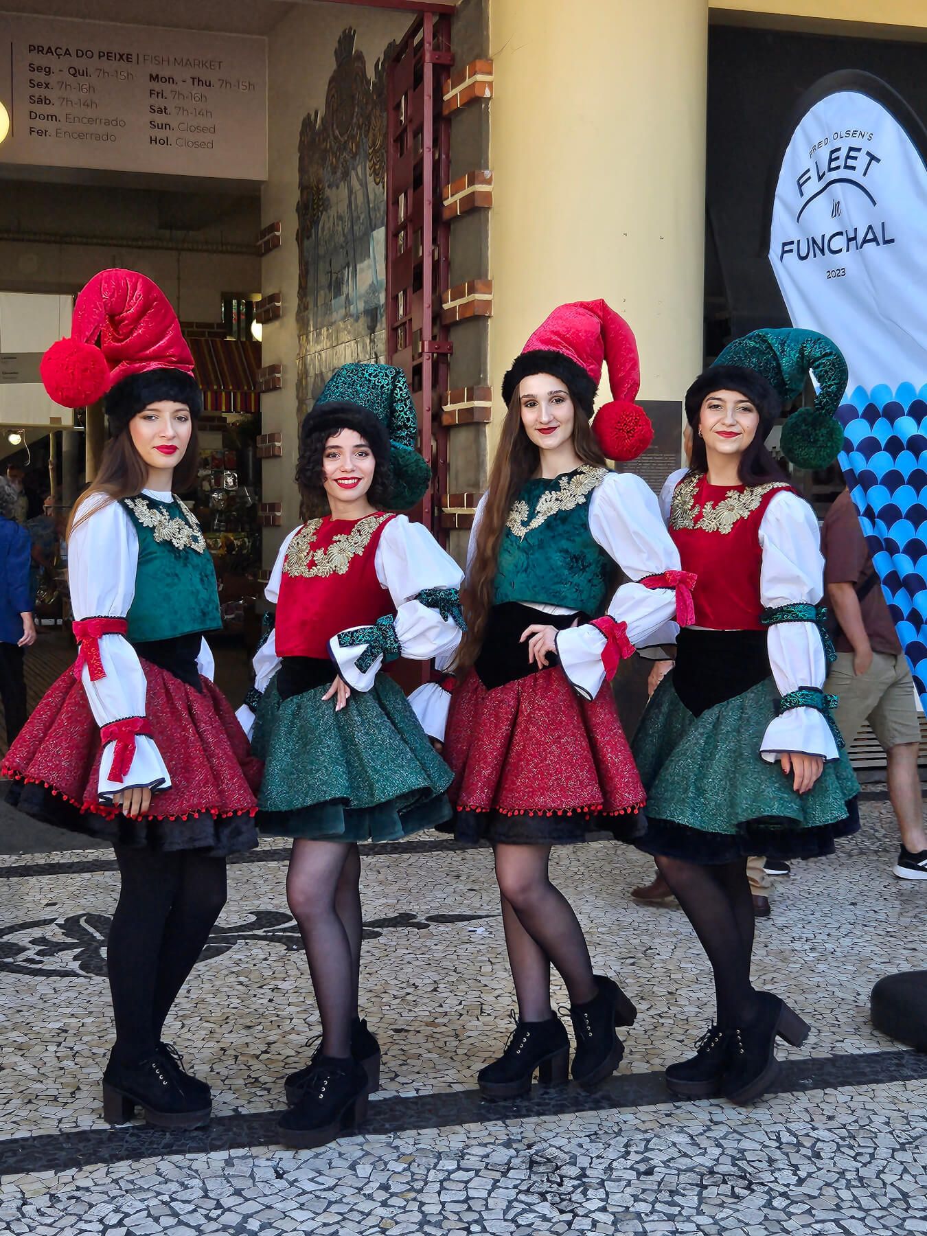 Vier Frauen in bunten Trachten mit großen Hüten posieren fröhlich vor einem Markt.