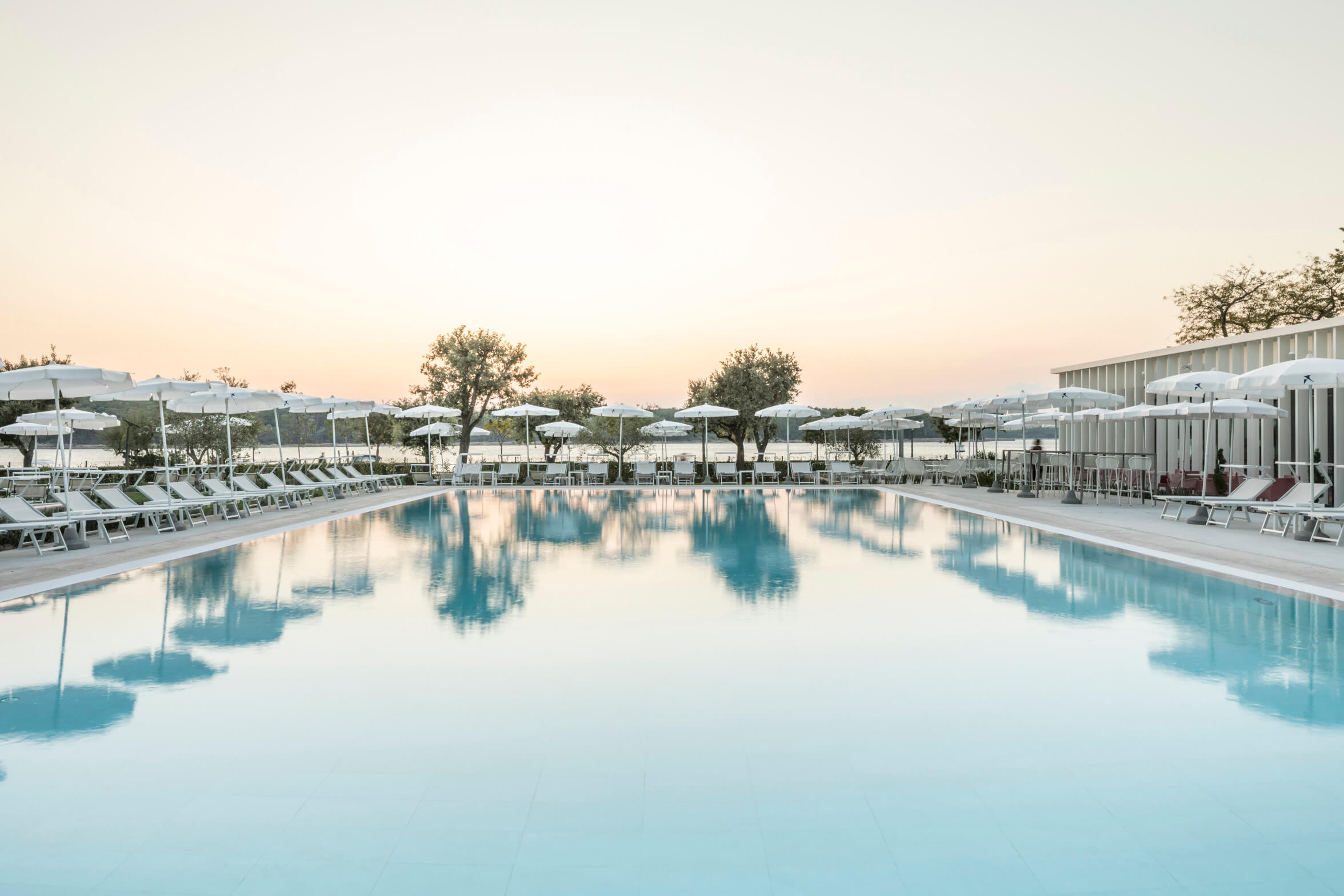 Ein ruhiger Pool mit Liegen und Sonnenschirmen, umgeben von Bäumen, bei Sonnenuntergang.