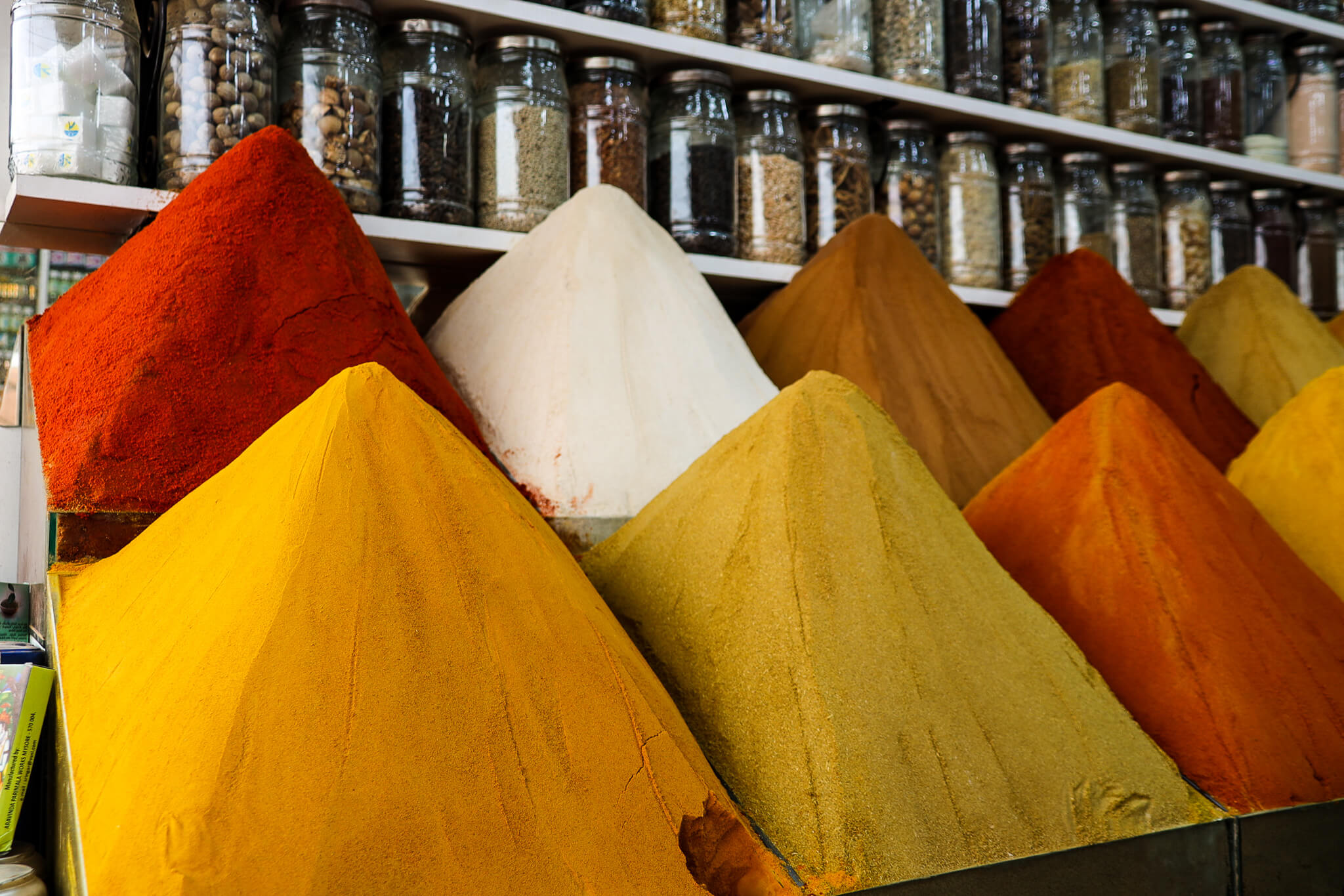 Bunte Gewürzhaufen in einer Ladenauslage, umgeben von Gläsern mit verschiedenen Zutaten.