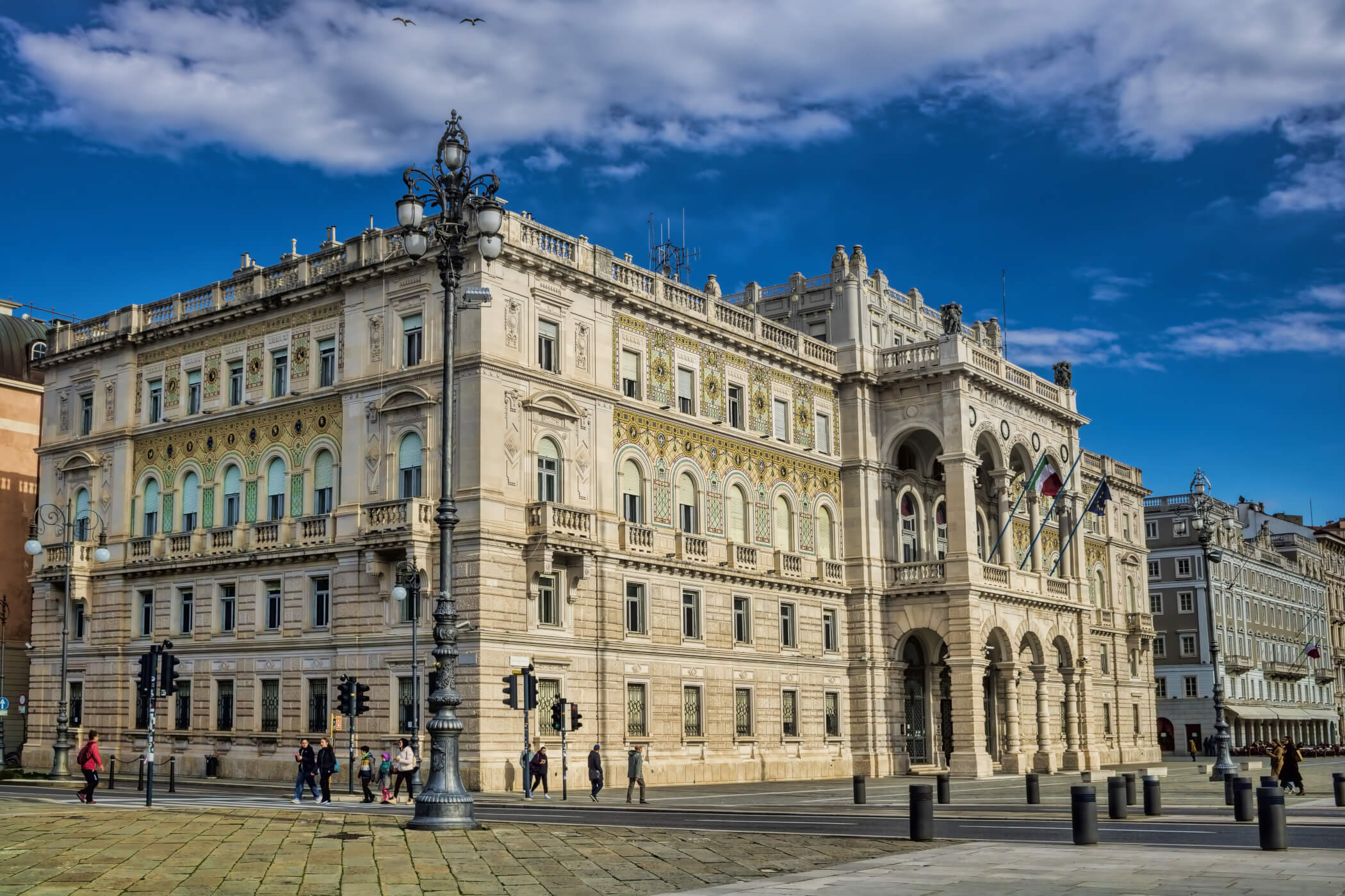 Ein prächtiges historisches Gebäude mit kunstvollen Details unter blauem Himmel und Wolken. Spaziergänger sind zu sehen.