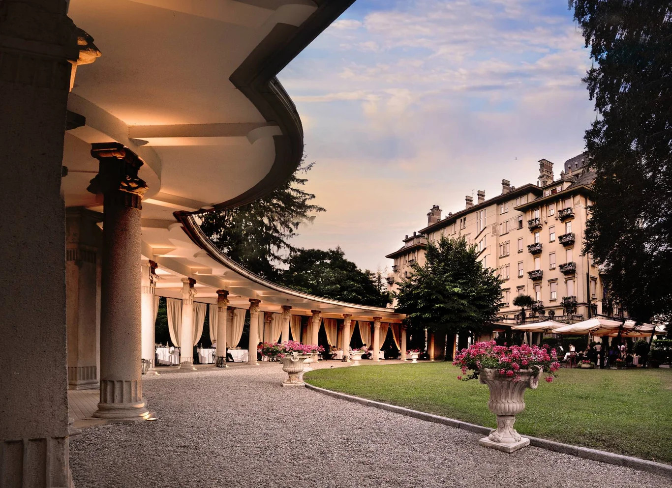 Ein eleganter Garten mit Säulen, Blumen und einem historischen Gebäude im Hintergrund bei Sonnenuntergang.