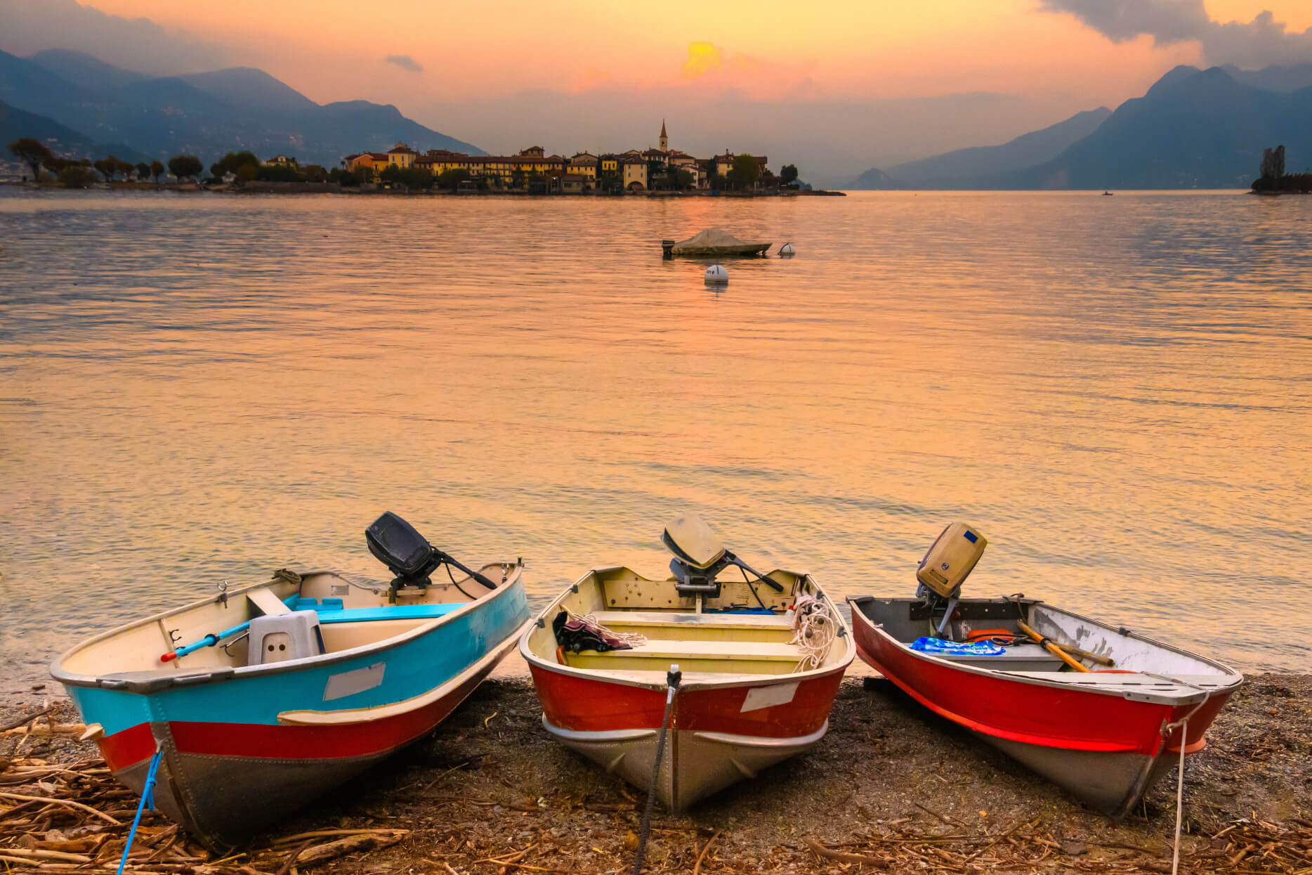 Drei Boote am Ufer, im Hintergrund eine malerische Stadt bei Sonnenuntergang und sanften Bergen.