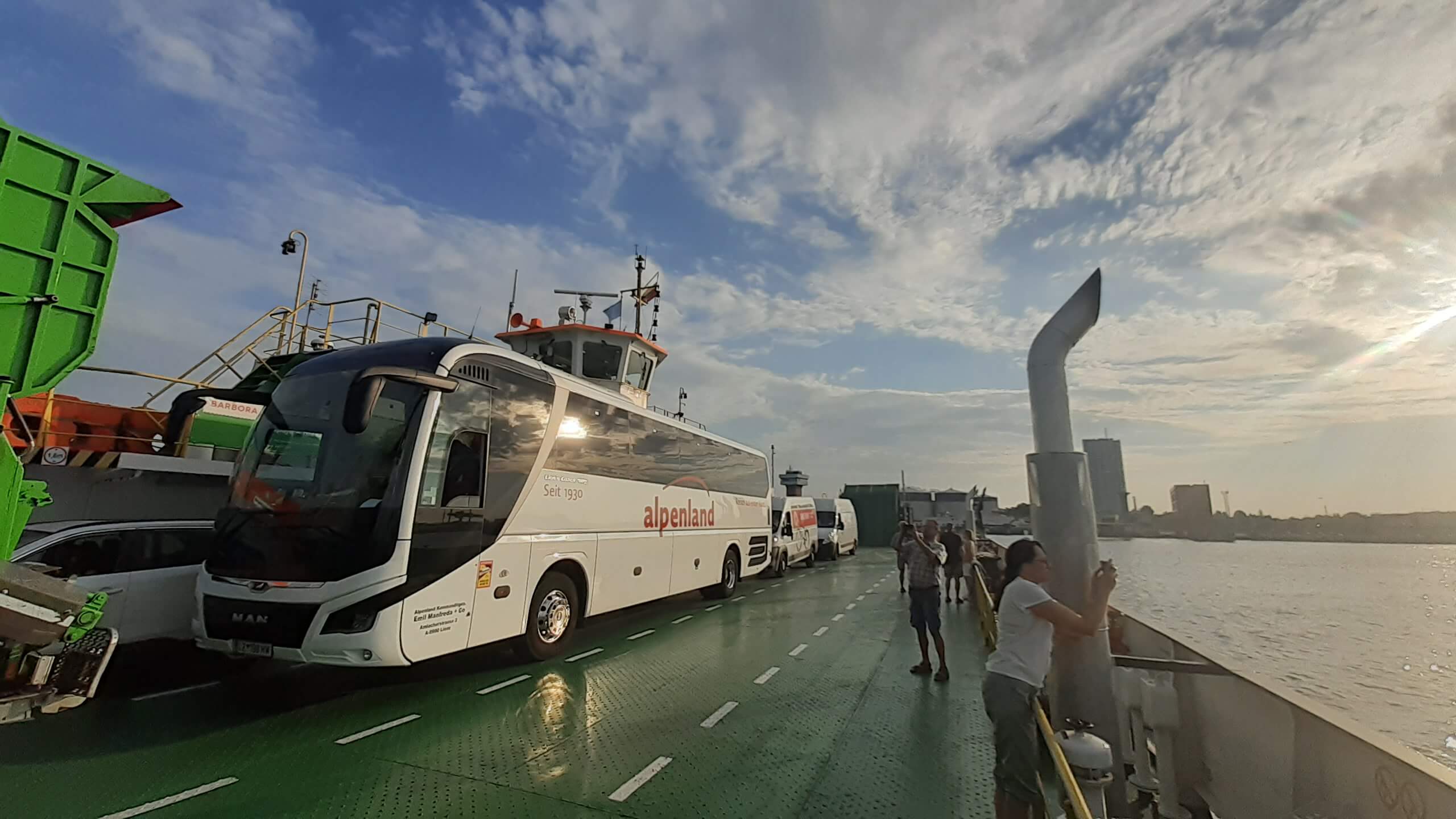 Ein Bus auf einem Schiff, Passagiere genießen die Aussicht, Wolken und Sonne am Himmel.