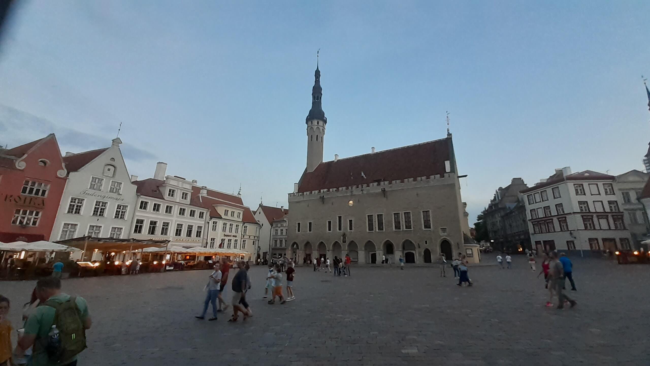 Ein Platz mit historischen Gebäuden und einem hohen Turm, in lebhafter Abendstimmung mit vielen Passanten.