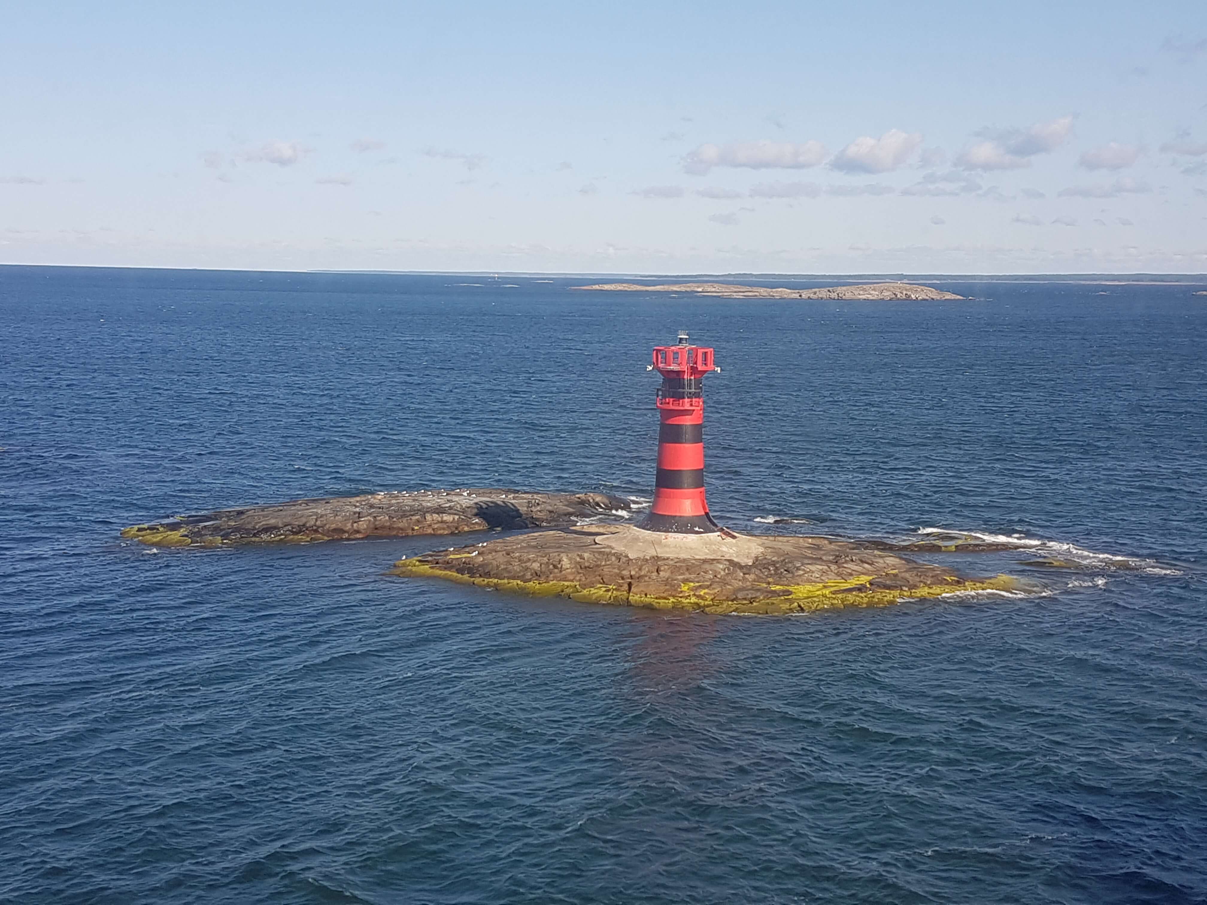 Ein roter und schwarzer Leuchtturm auf einer kleinen Felseninsel im blauen Meer unter einem klaren Himmel.