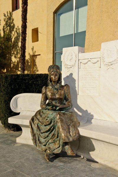 Eine Bronzeskulptur einer Dame in historischem Kleid sitzt auf einer marmornen Bank vor einer Wand.