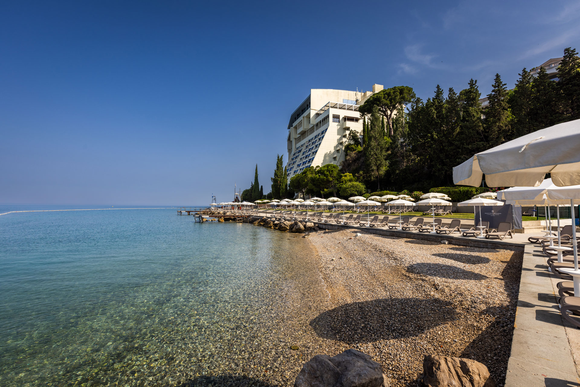 Ein ruhiger Strand mit klarem Wasser, Liegen und Schirmen, umgeben von Bäumen und einem modernen Gebäude.