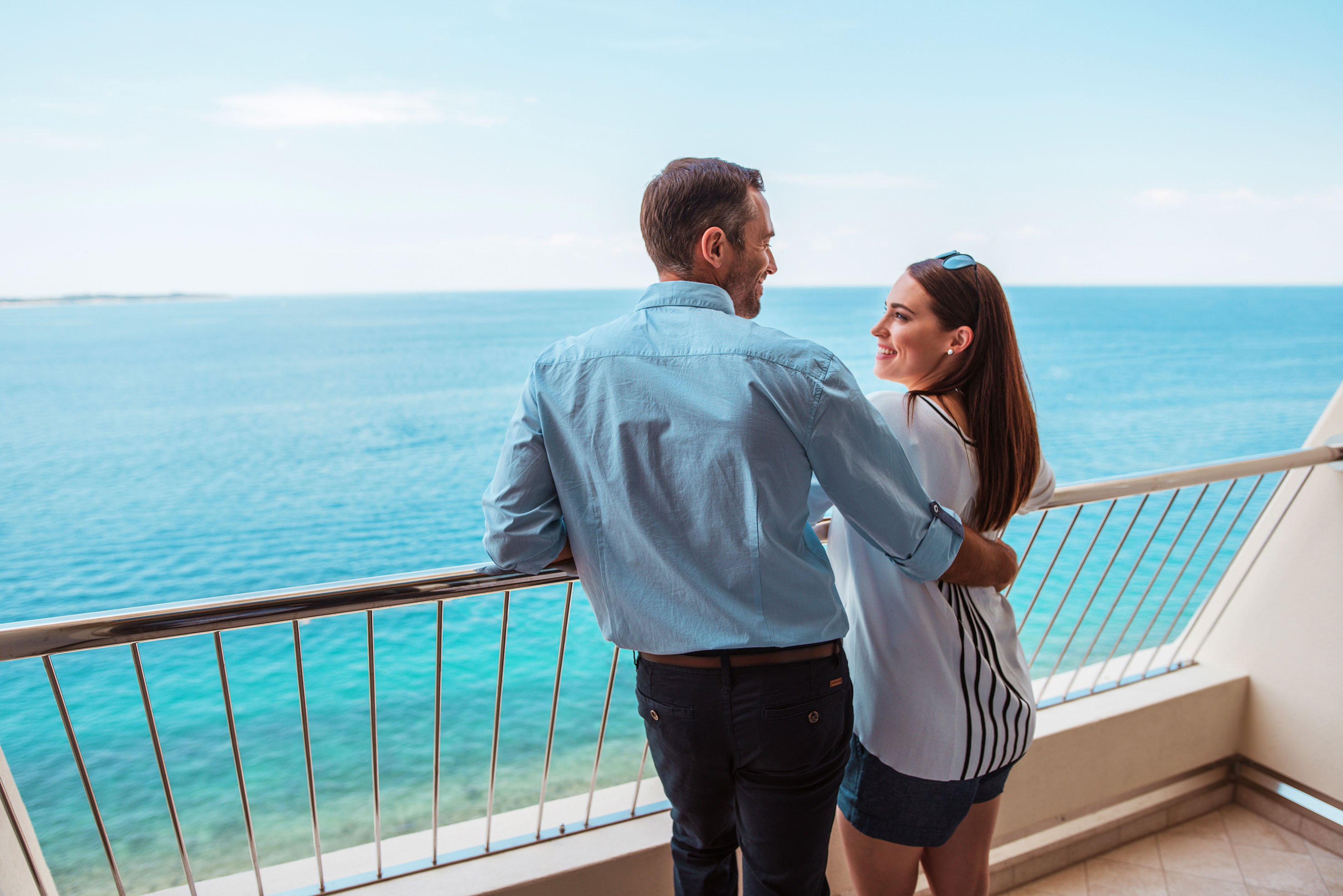 Ein Paar steht am Balkon und genießt den Blick auf das ruhige blaue Meer. Ein romantischer Moment.