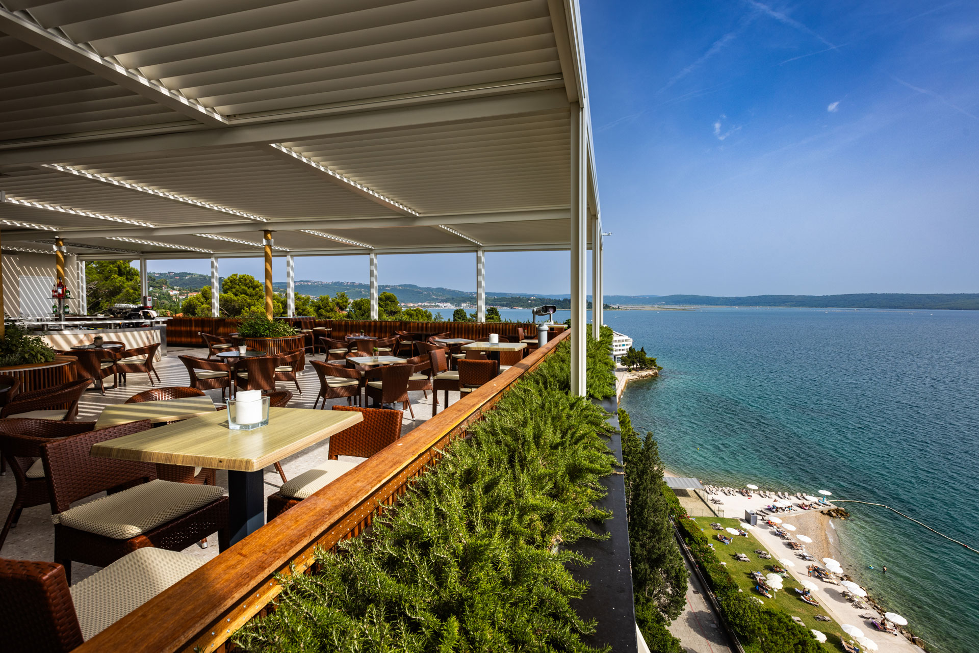 Terrasse eines Restaurants mit Blick auf das Wasser, komfortable Stühle und Tische unter einem modernen Dach.