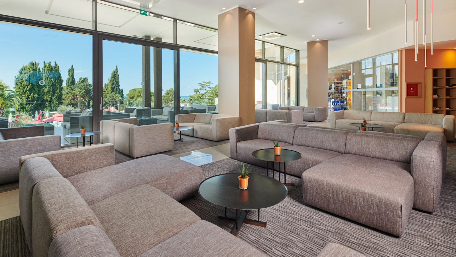 Moderne Lounge mit bequemen Sofas, Tischen und Blick ins Freie auf Bäume und einen blauen Himmel.