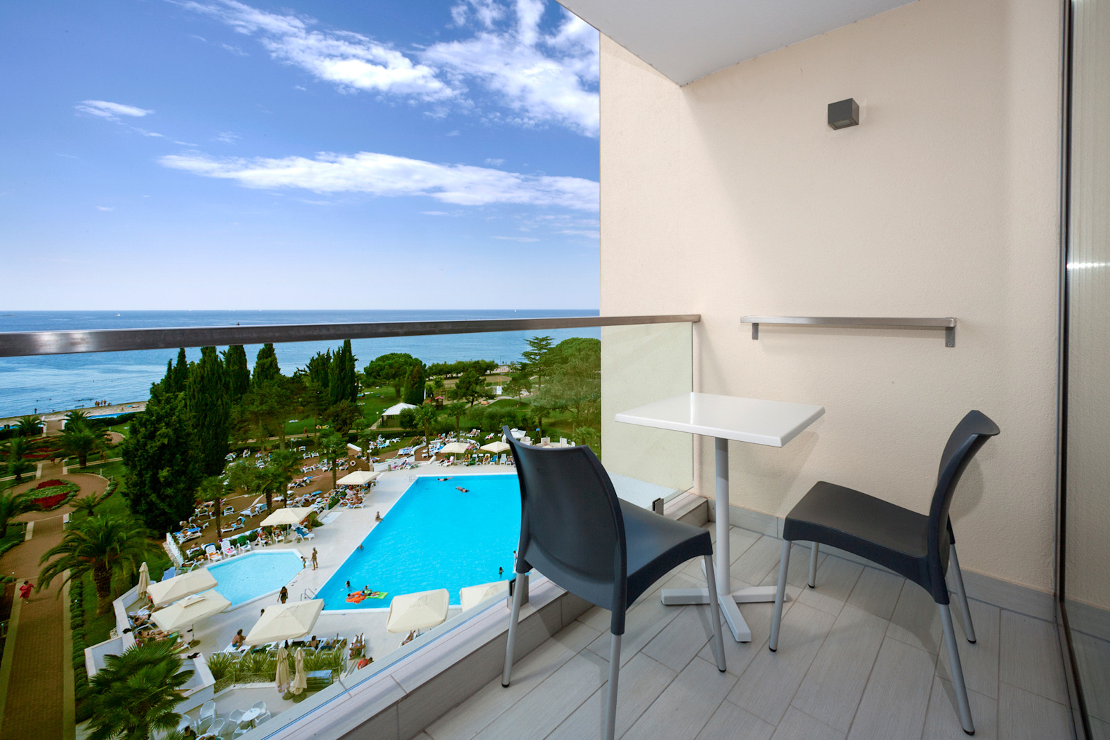 Balkon mit Blick auf den Pool und das Meer, moderne Sitzecke, sonniger Tag mit klarem Himmel.