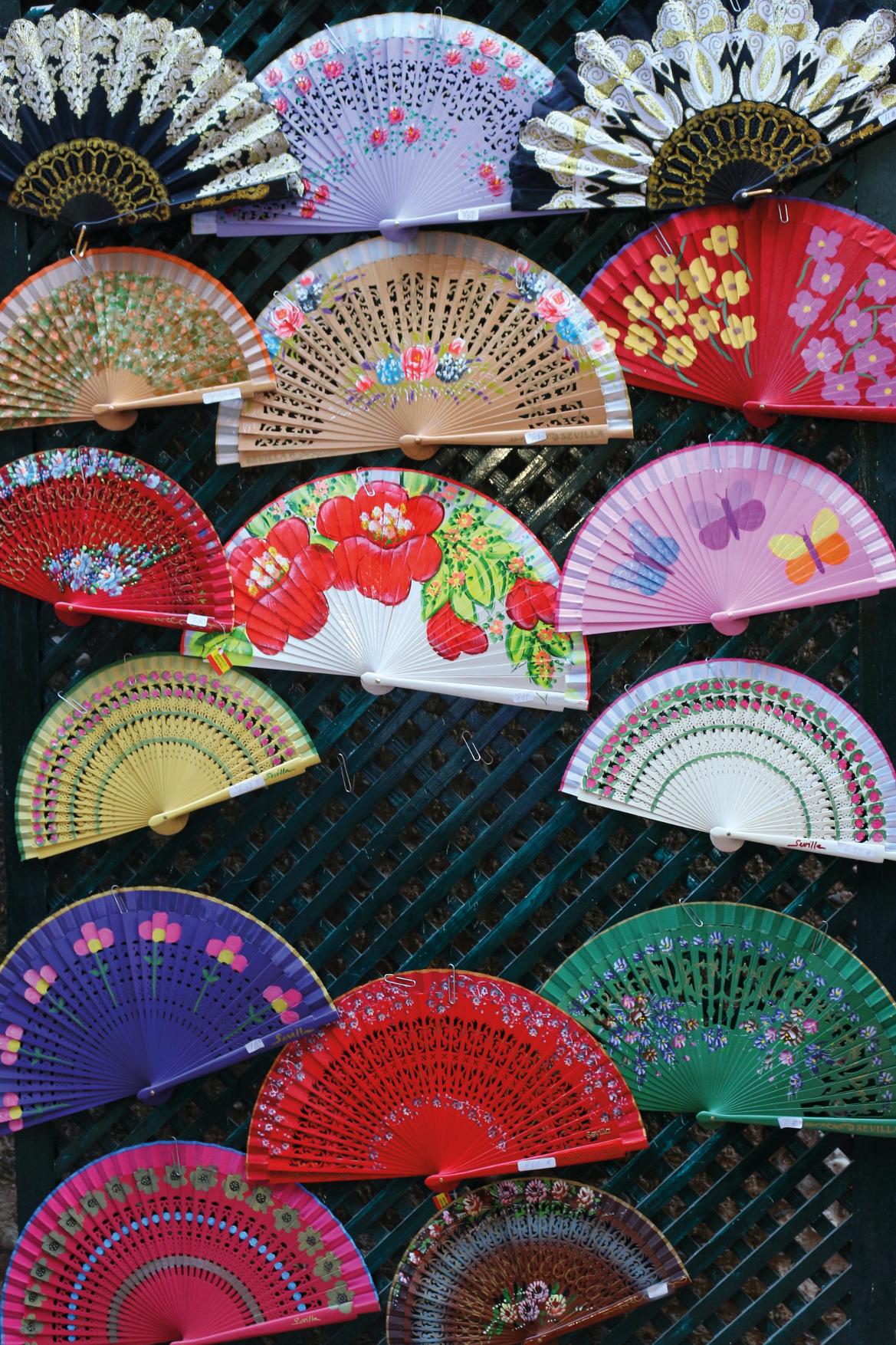 Eine bunte Sammlung handgefertigter Fächer, die an einem Gitter aufgehängt sind. Vielfalt in Design und Farben.