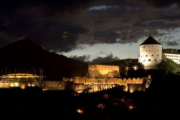 Eine beleuchtete Burg steht majestätisch im Nachtlicht, umgeben von Bergen und dramatischen Wolken.