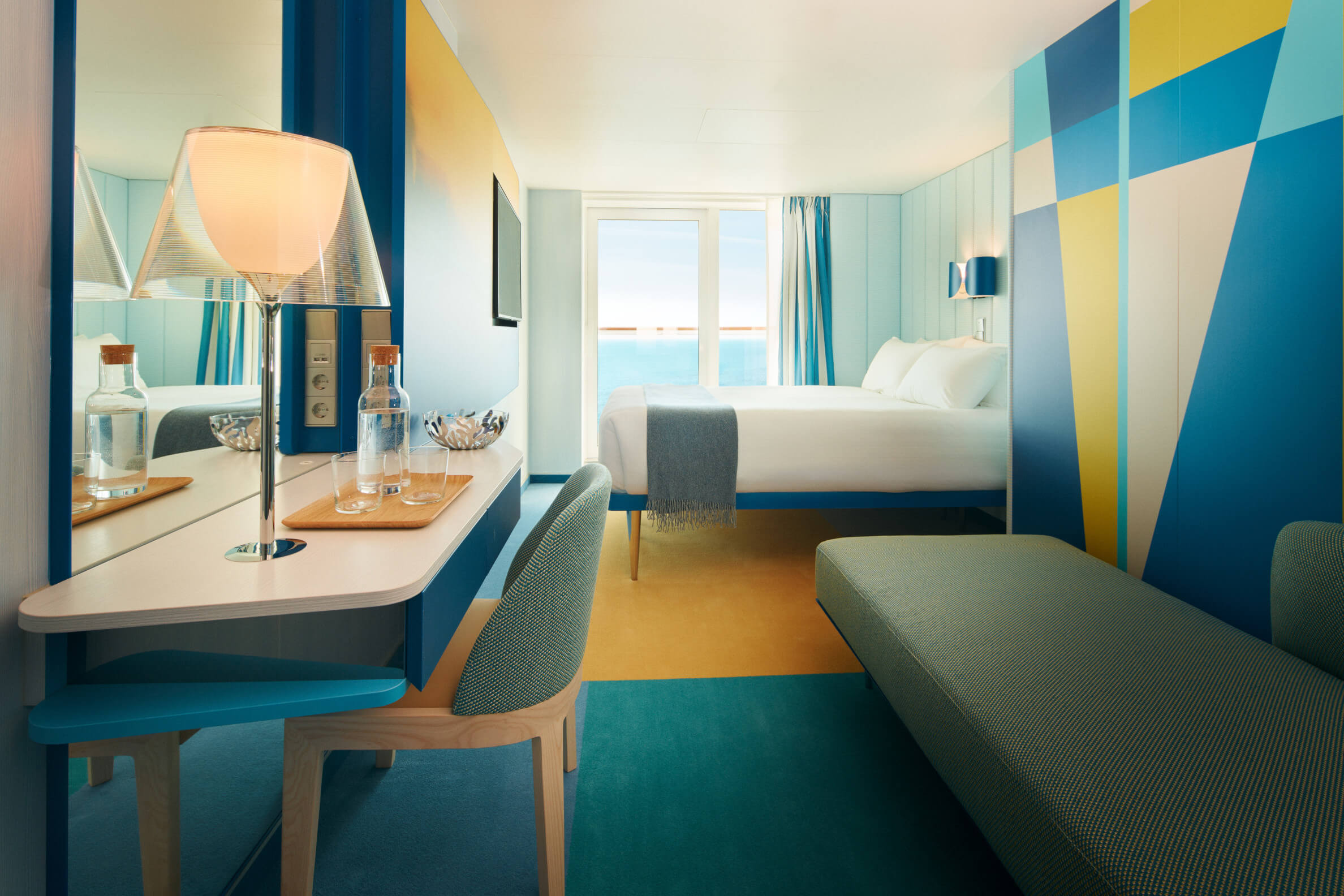 Modernes Hotelzimmer in Blau- und Gelbtönen mit Blick auf das Meer, ausgestattet mit einem Schreibtisch und Lounge-Bereich.