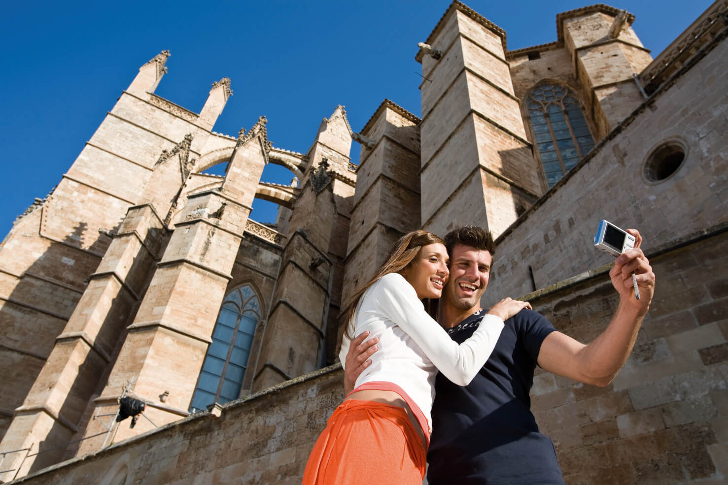 Ein Paar macht ein Selfie vor einer beeindruckenden Kathedrale unter blauem Himmel. Sie strahlen Freude aus.