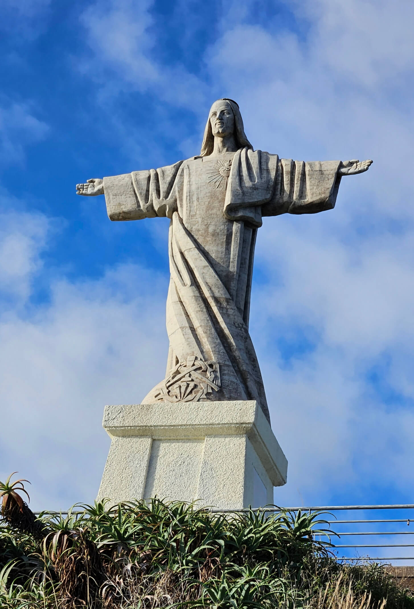 Eine majestätische Christusstatue mit ausgebreiteten Armen vor einem blauen Himmel und üppigem Grün im Vordergrund.