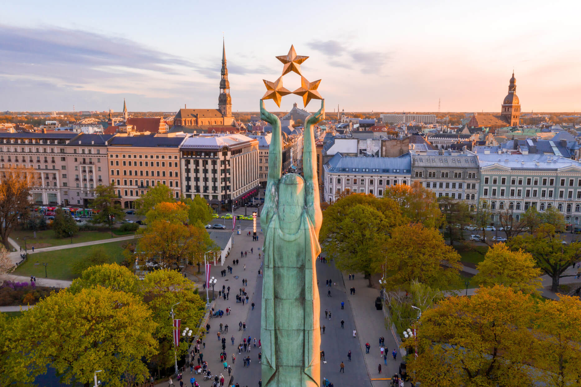 Ein Blick auf die Freiheitsstatue in Riga mit historischem Stadtbild und Bäumen im Vordergrund.