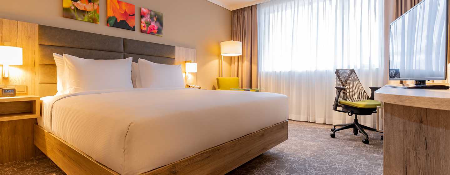 Ein modernes Hotelzimmer mit einem großen Bett, Schreibtisch, Fernseher und hellem Fenster. Dekorative Wandbilder sind sichtbar.