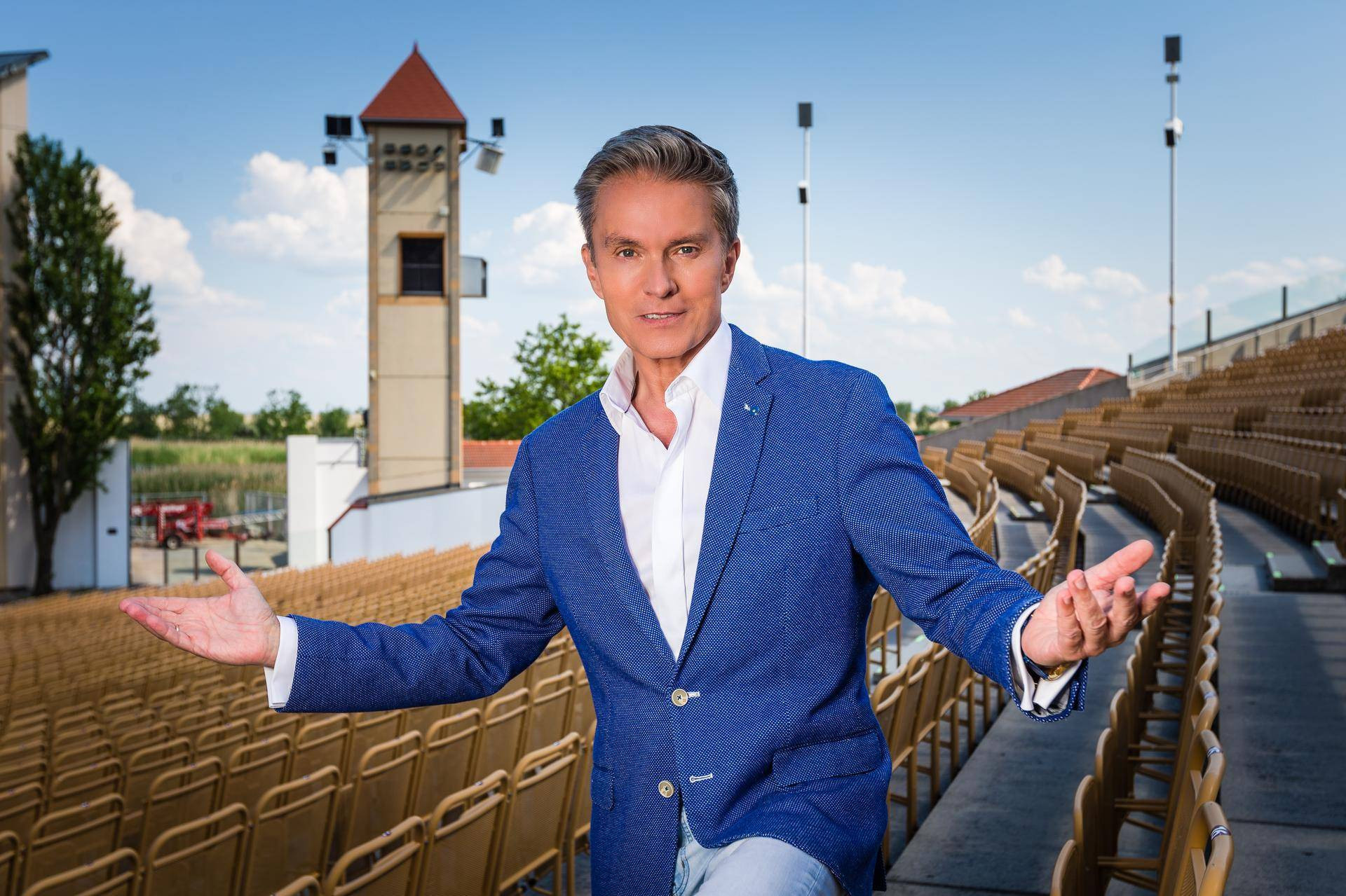 Ein Mann in einem blauen Blazer steht auf einer leeren Freiluftbühne und begrüßt das Publikum.