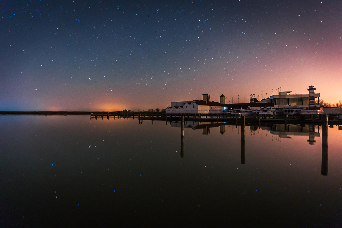 Ein ruhiger Hafen bei Nacht, Sterne spiegeln sich im Wasser, sanfte Farben am Horizont.