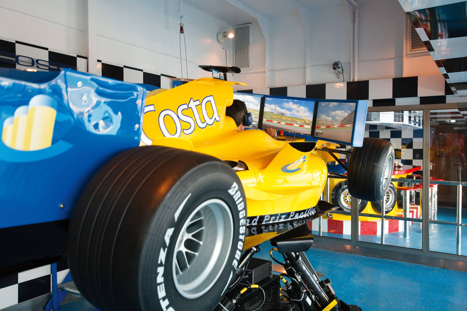 Ein gelbes Formel-1-Auto mit zwei Bildschirmen steht in einem Raum mit Motorsport-Dekor.