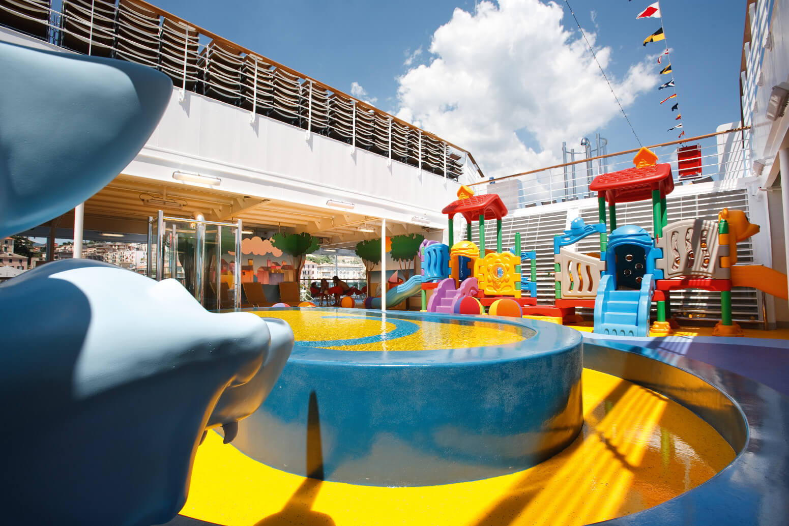 Ein bunter Spielplatz auf einem Schiff mit Wasserbecken und verschiedenen Spielgeräten. Heller Himmel im Hintergrund.