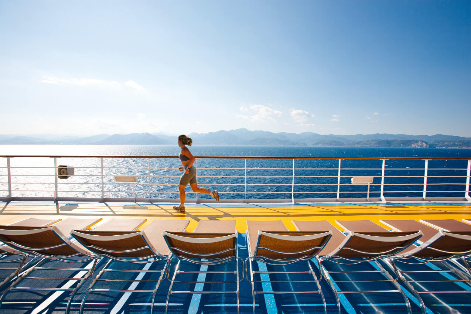 Eine Frau joggt auf dem Deck eines Kreuzfahrtschiffs mit Blick auf das Meer und die Berge im Hintergrund.