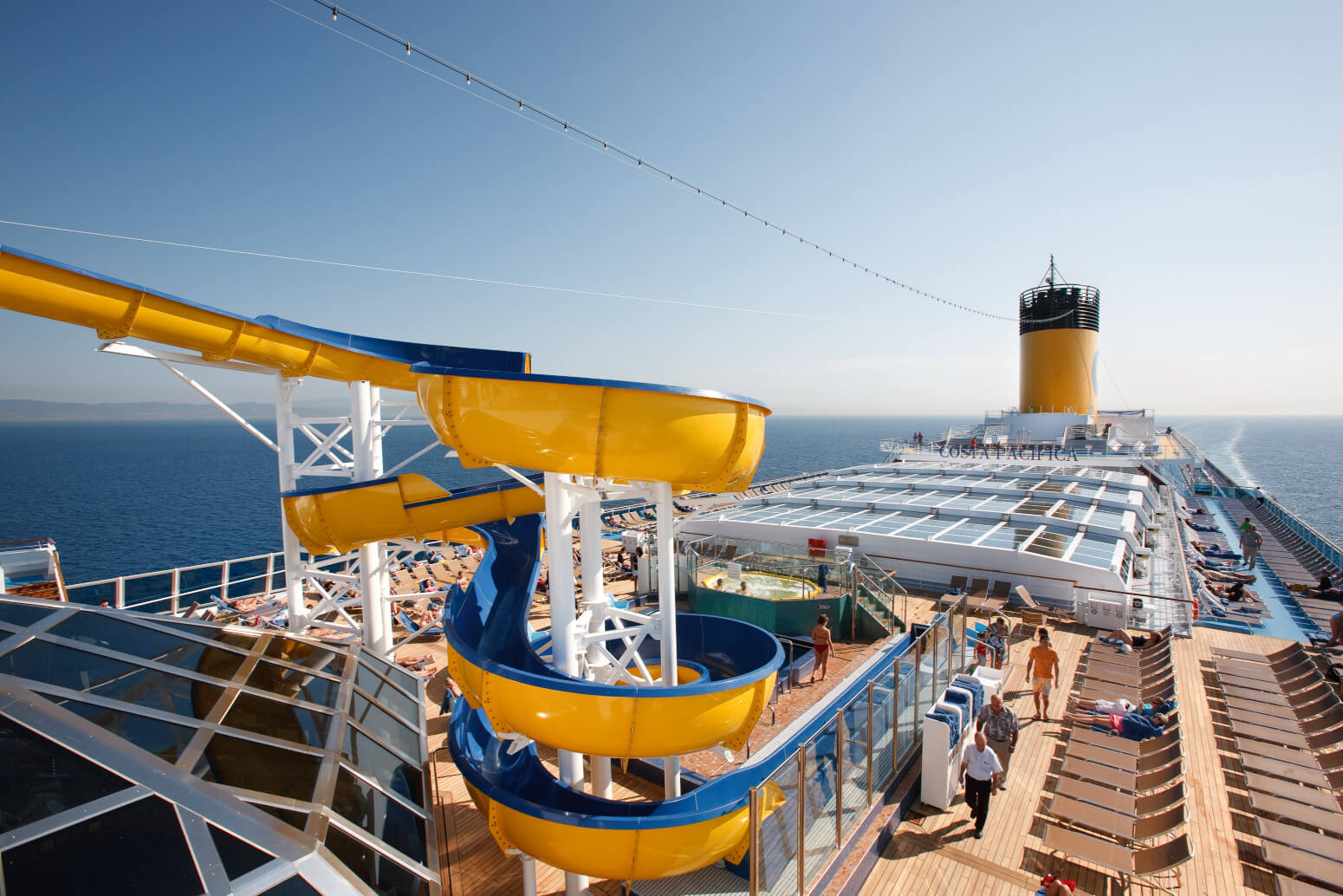 Ein Kreuzfahrtschiff mit einer gelben Wasserrutsche und Sonnenliegen, umgeben von Meer und blauem Himmel.