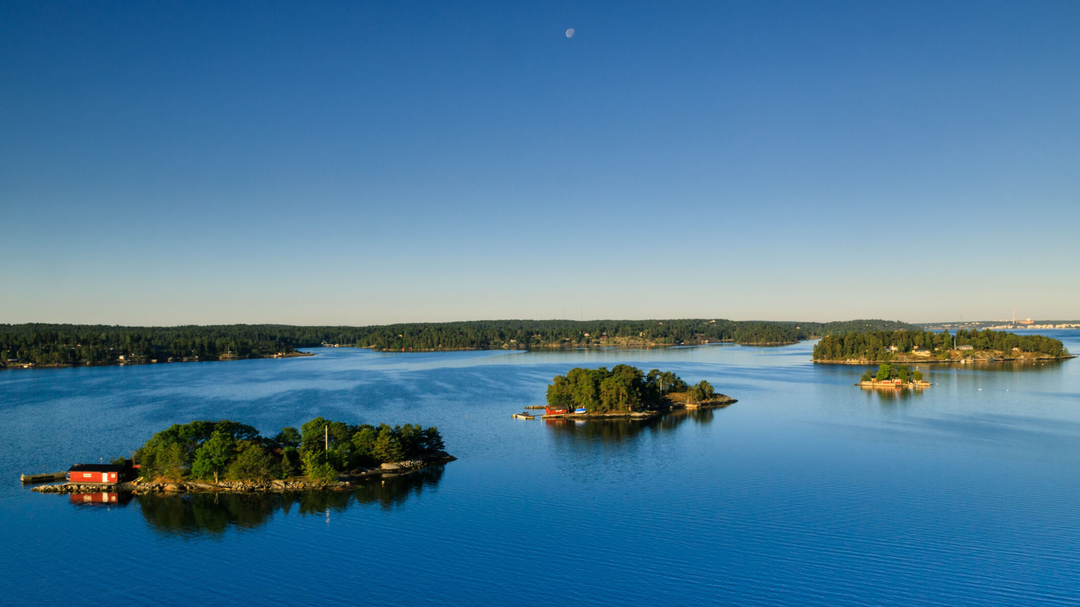 Ein klarer blauer Himmel über ruhigem Wasser mit kleinen Inseln und Wäldern. Ruhe und Natur pur.