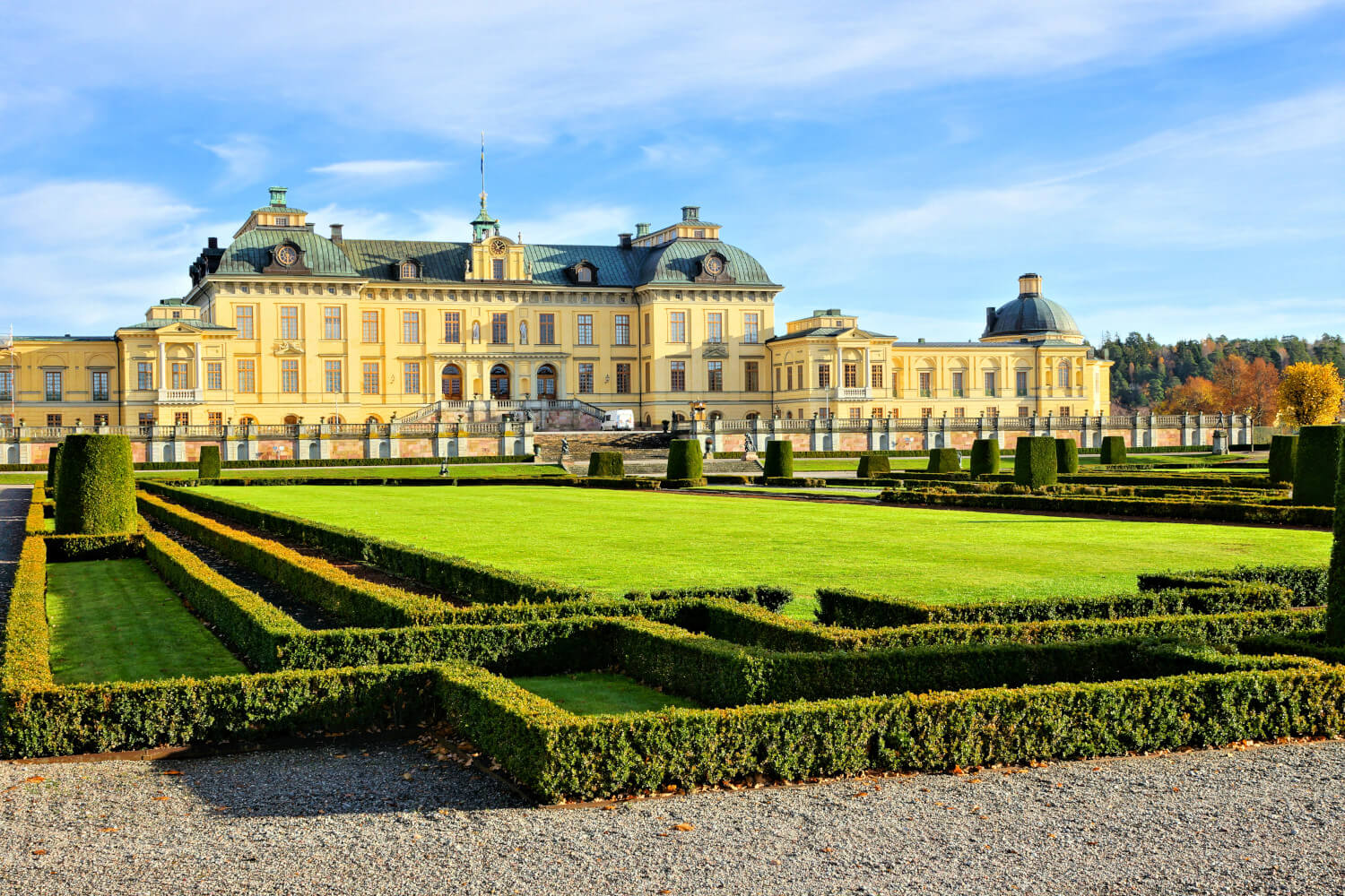 Ein prächtiges Schloss umgeben von kunstvoll gestalteten Gärten und gepflegtem Rasen im Sonnenlicht.
